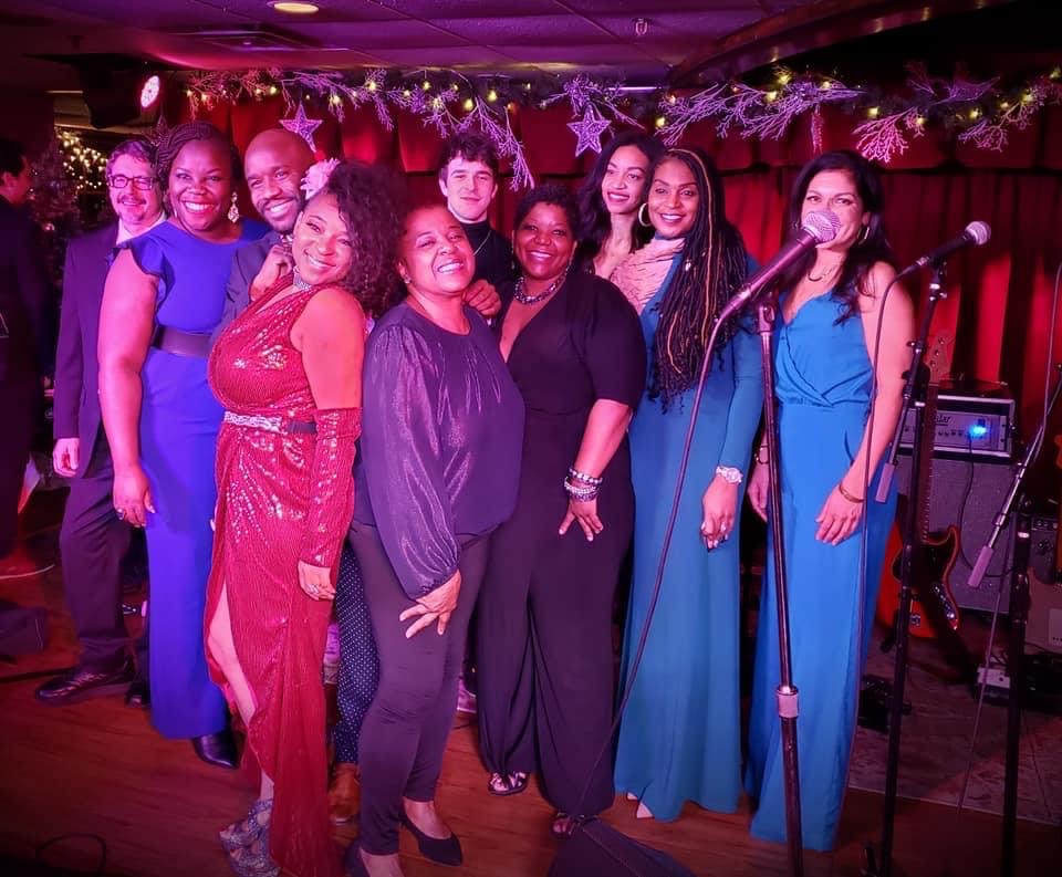 Group of ten people on stage, smiling, with festive decorations and a microphone, in a performance setting.