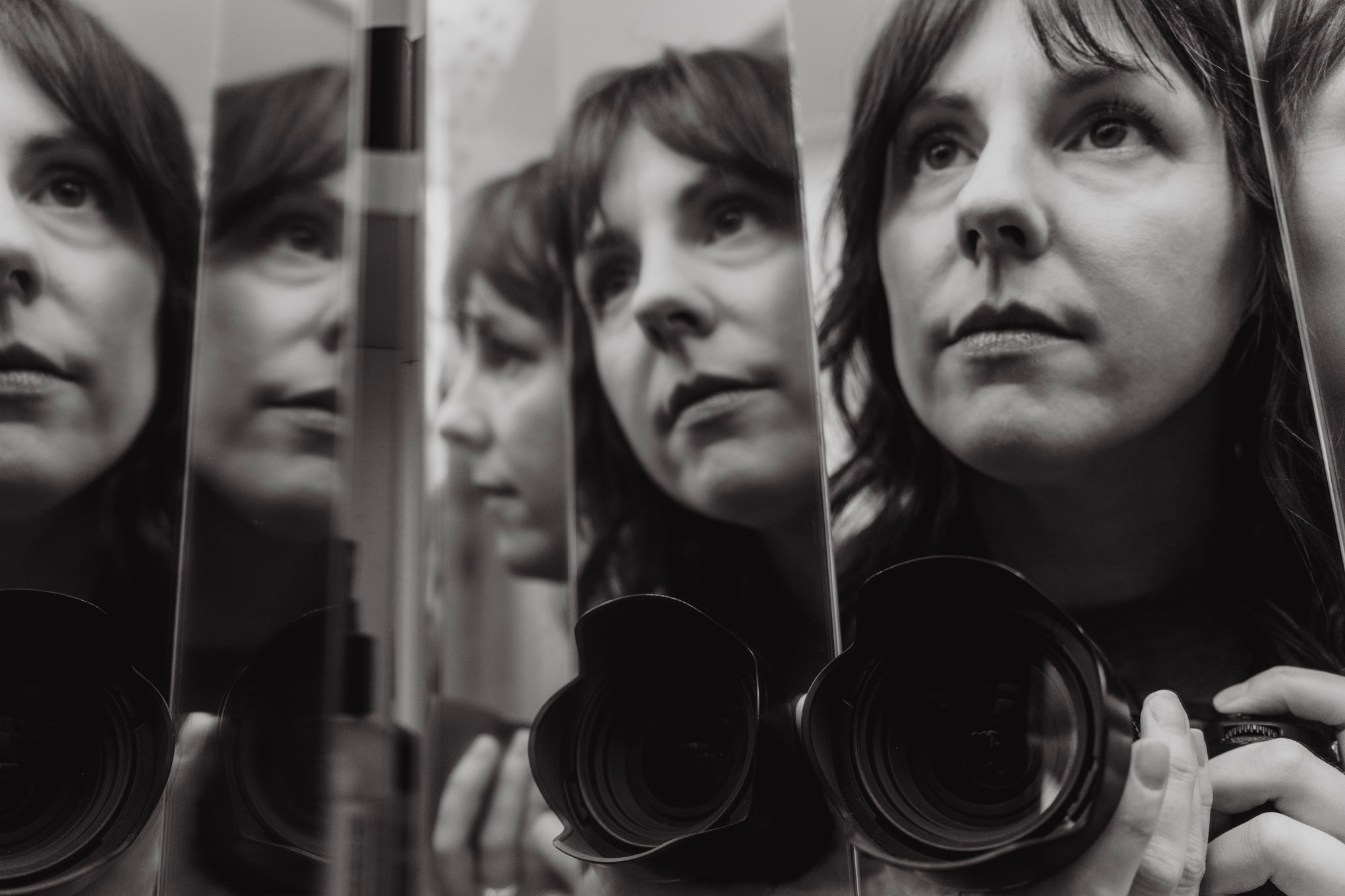 A woman looks at her reflection in multiple mirrors, capturing her face from different angles, with a camera in the foreground.