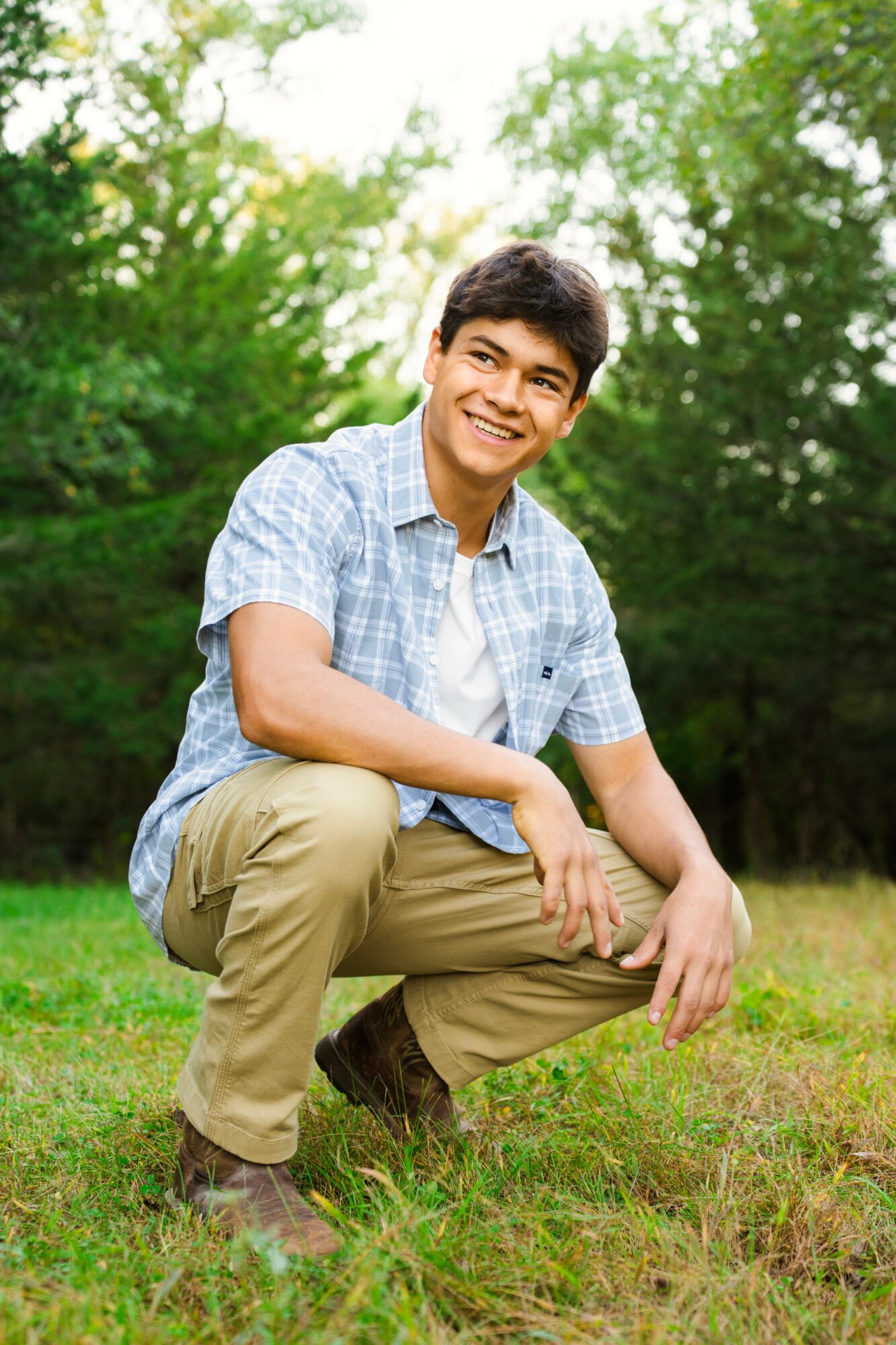 Young man crouching outdoors on grass, smiling, surrounded by green trees and bright sky.