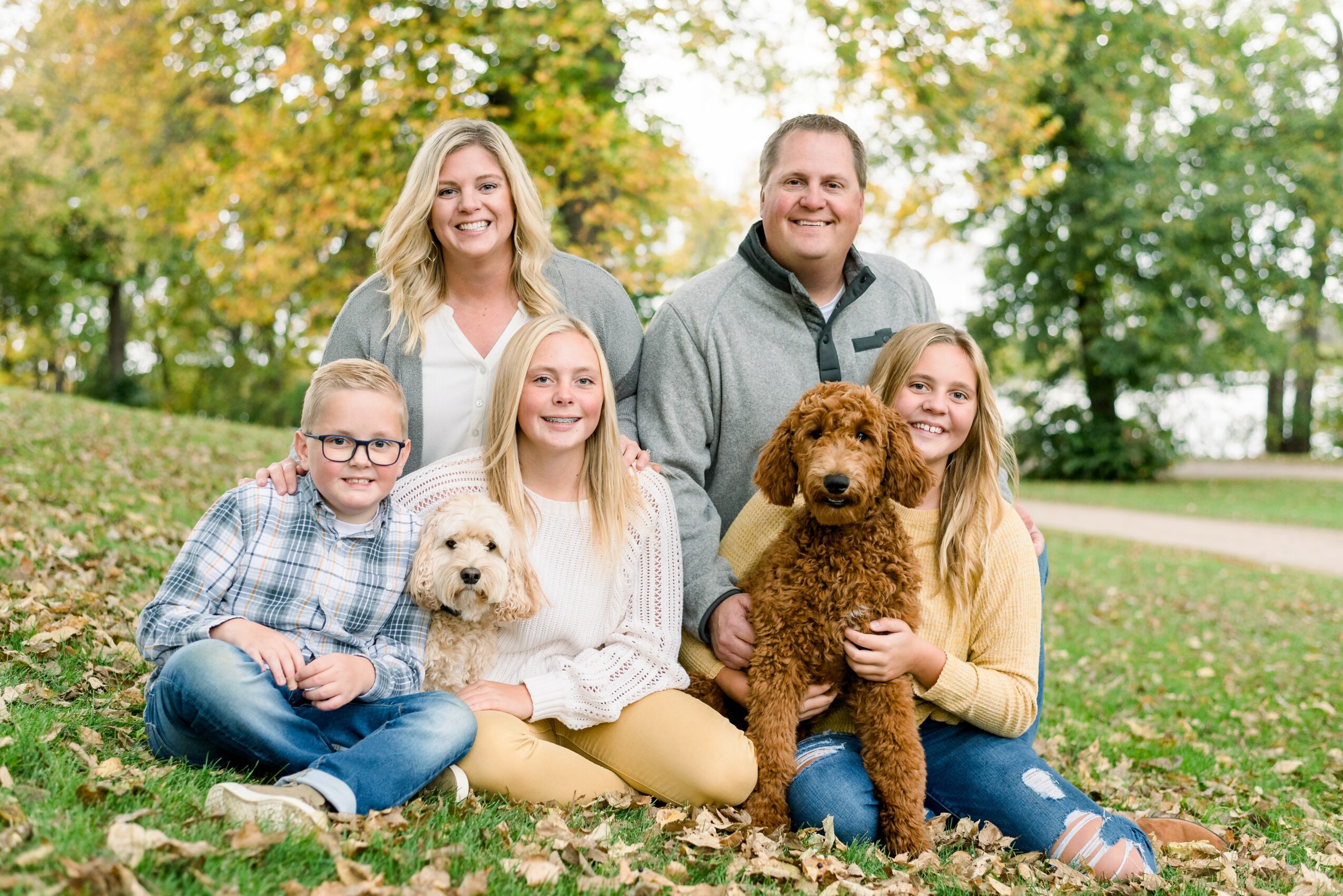 Family of five with two dogs sitting outdoors on grass, trees with autumn leaves in background.