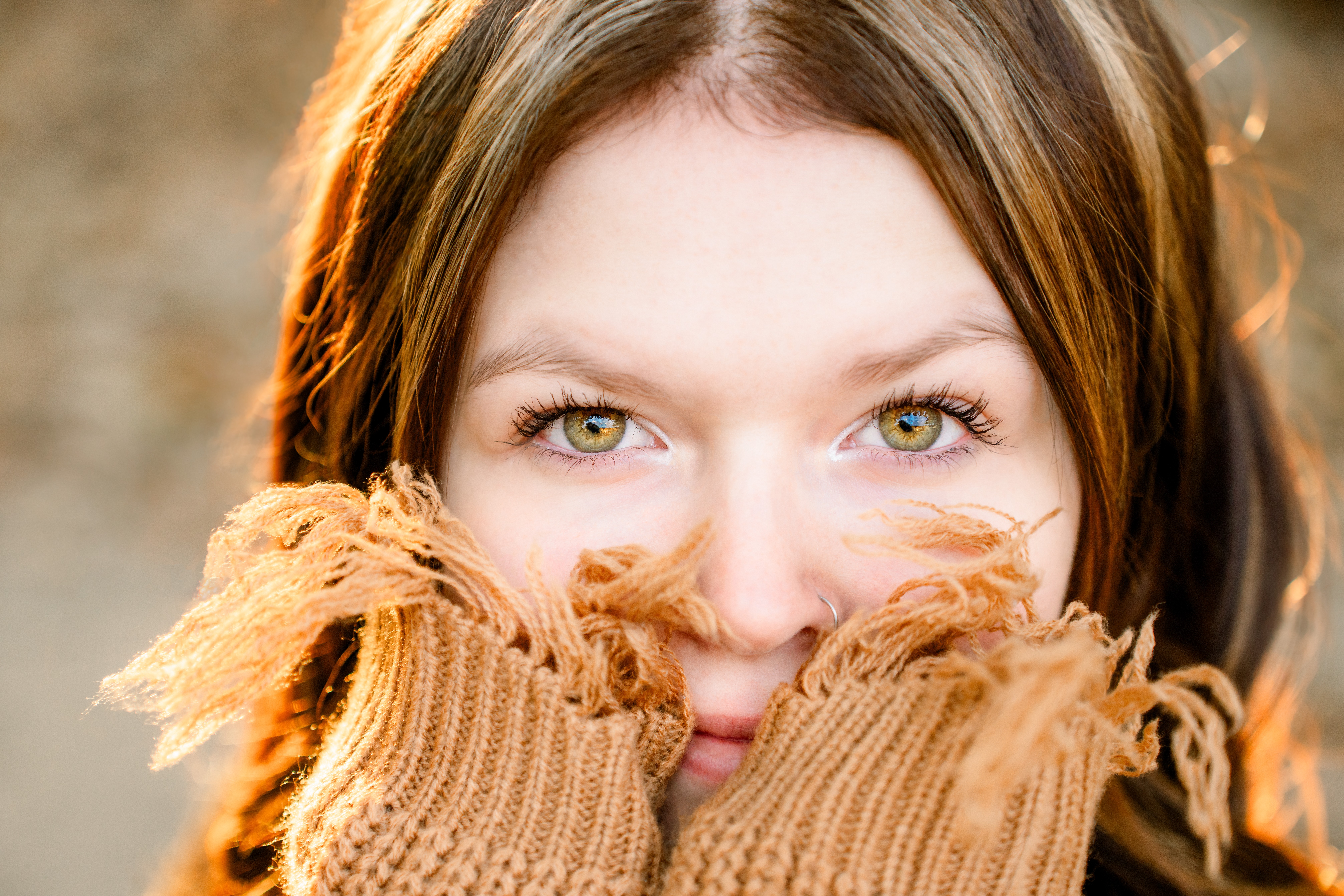 Close-up of a woman with light-colored eyes and brown hair, holding her sweater up to her face, outdoors.