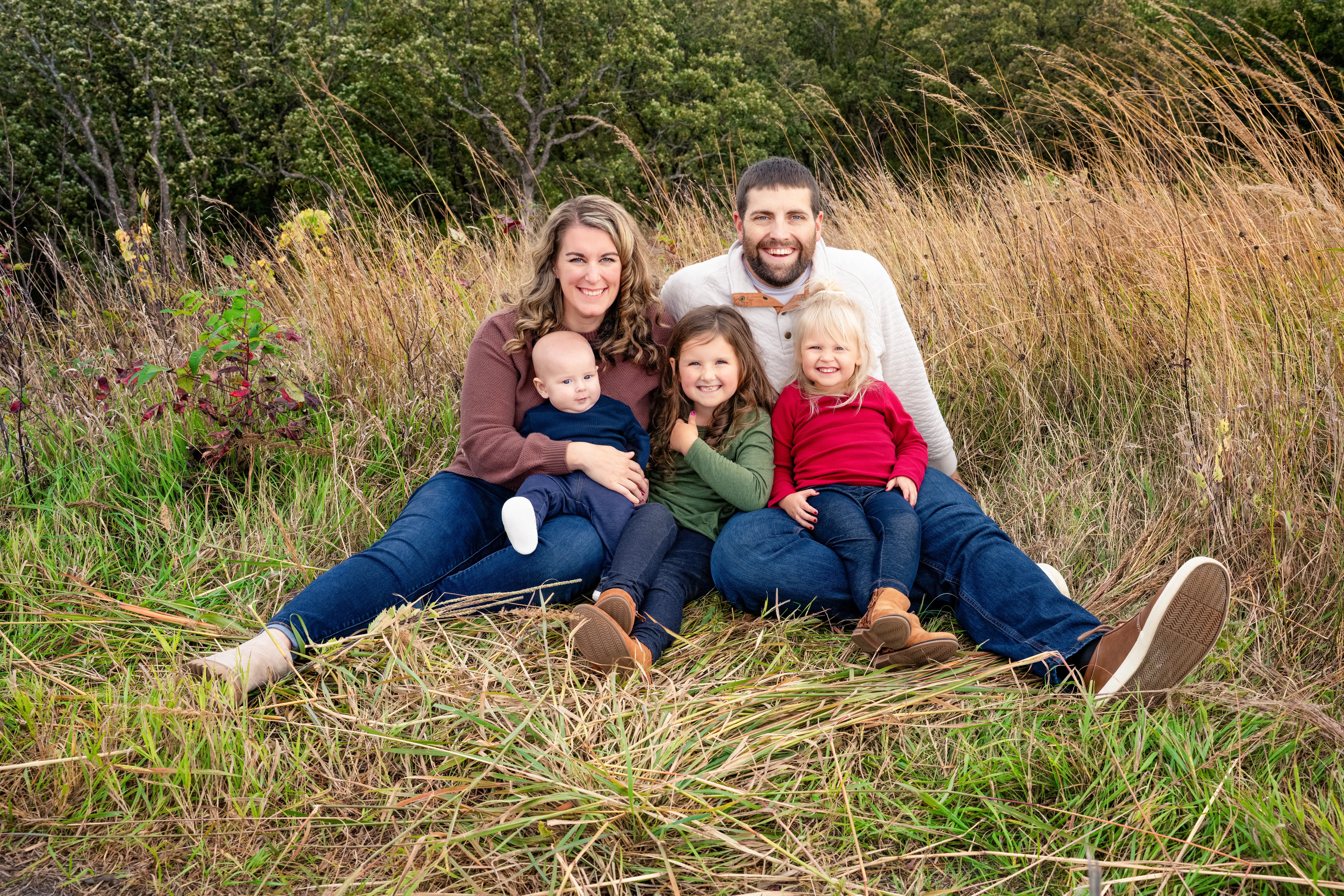 Family of five sitting on grass in a field, smiling, with trees and tall grass in background.