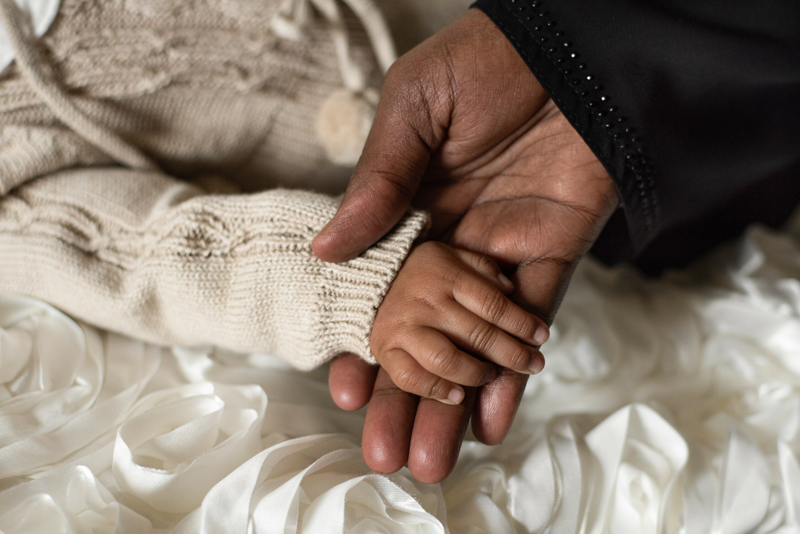 Adult hand holding a child's hand, both resting on a white fabric surface, child wearing a cream-colored sweater.