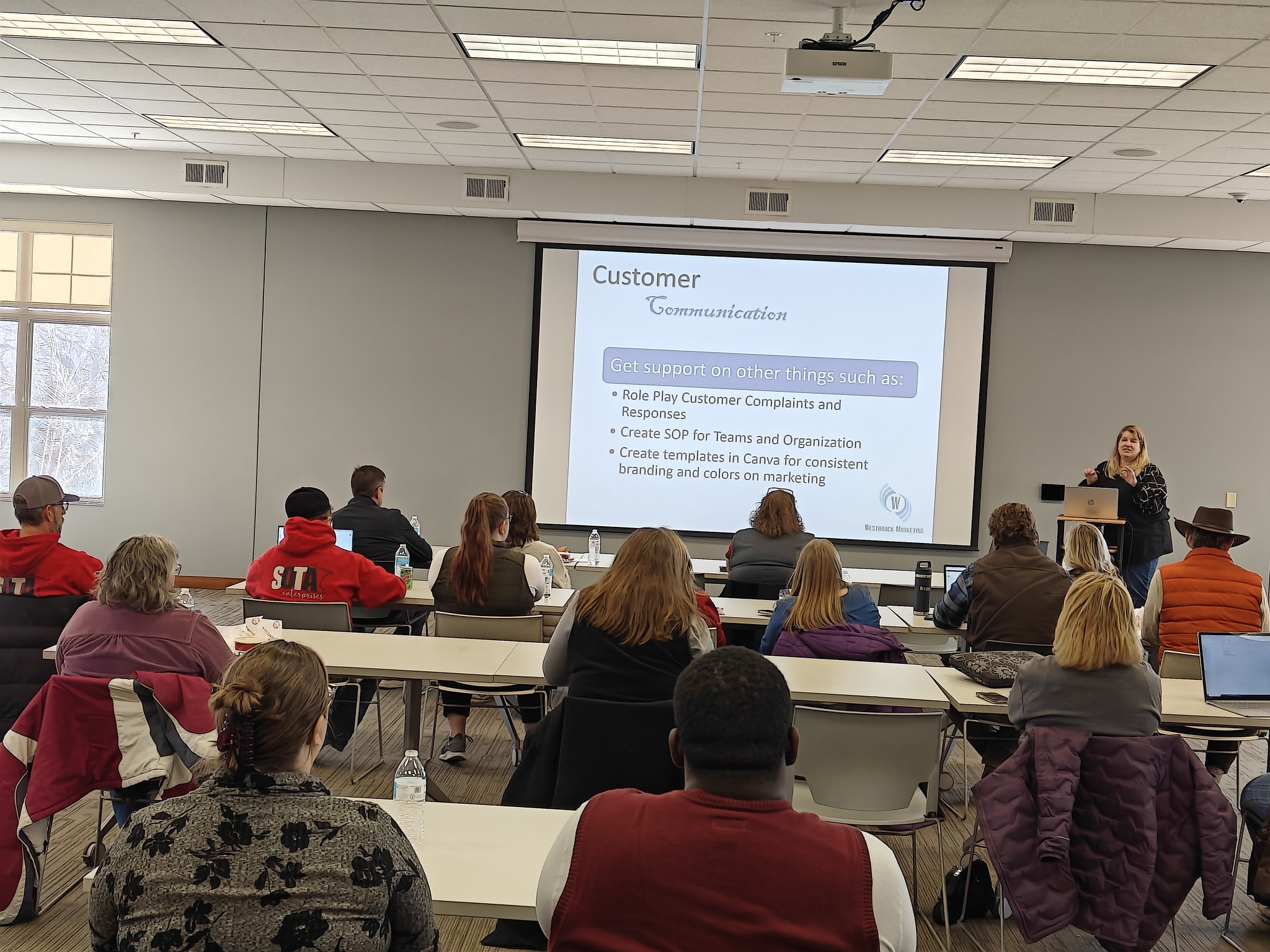 Classroom with people seated at tables, a presenter at a podium, and a presentation slide on the screen.