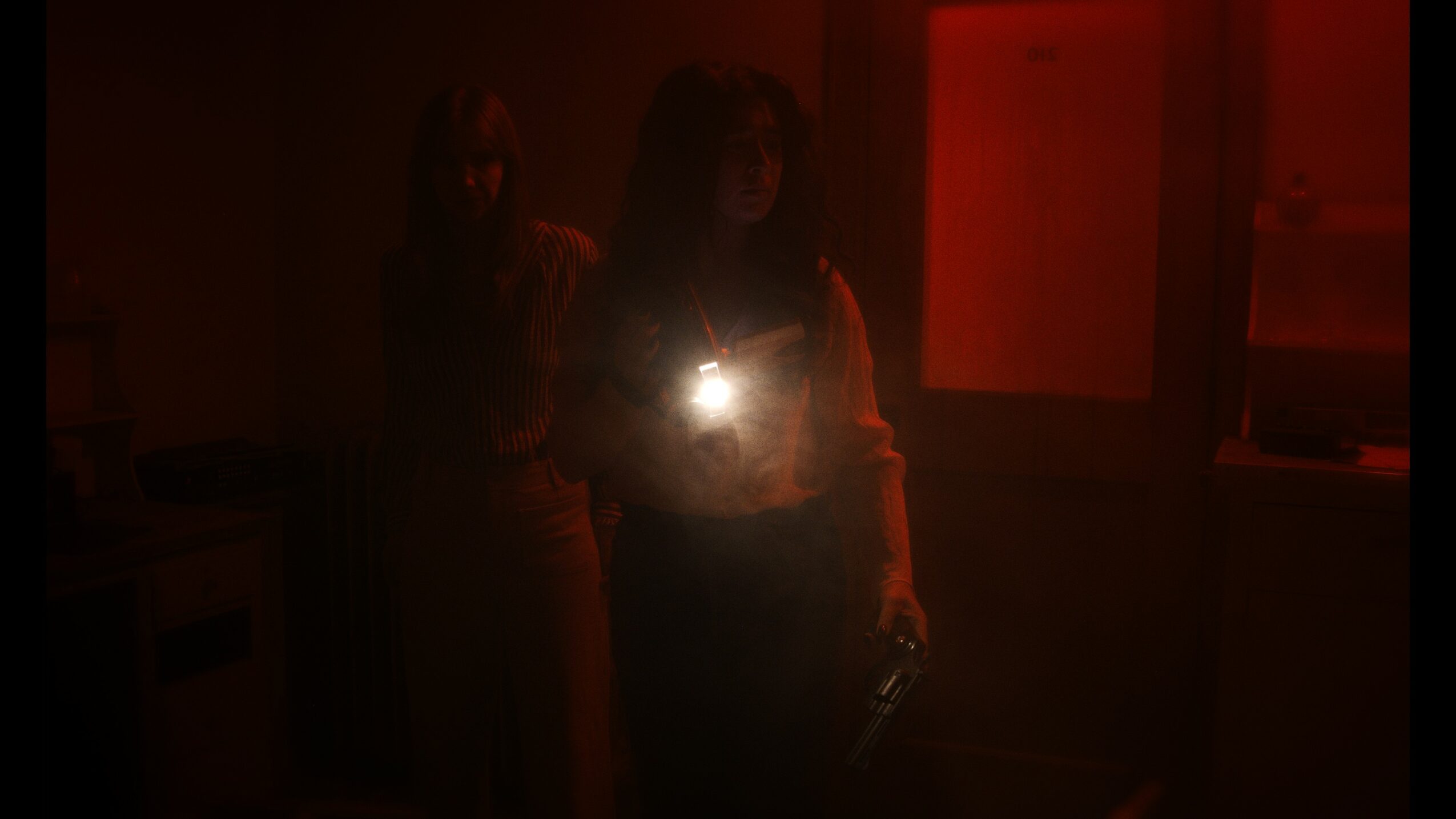 Two people standing in a dark room, one holding a phone with a bright flash, faintly visible background elements.