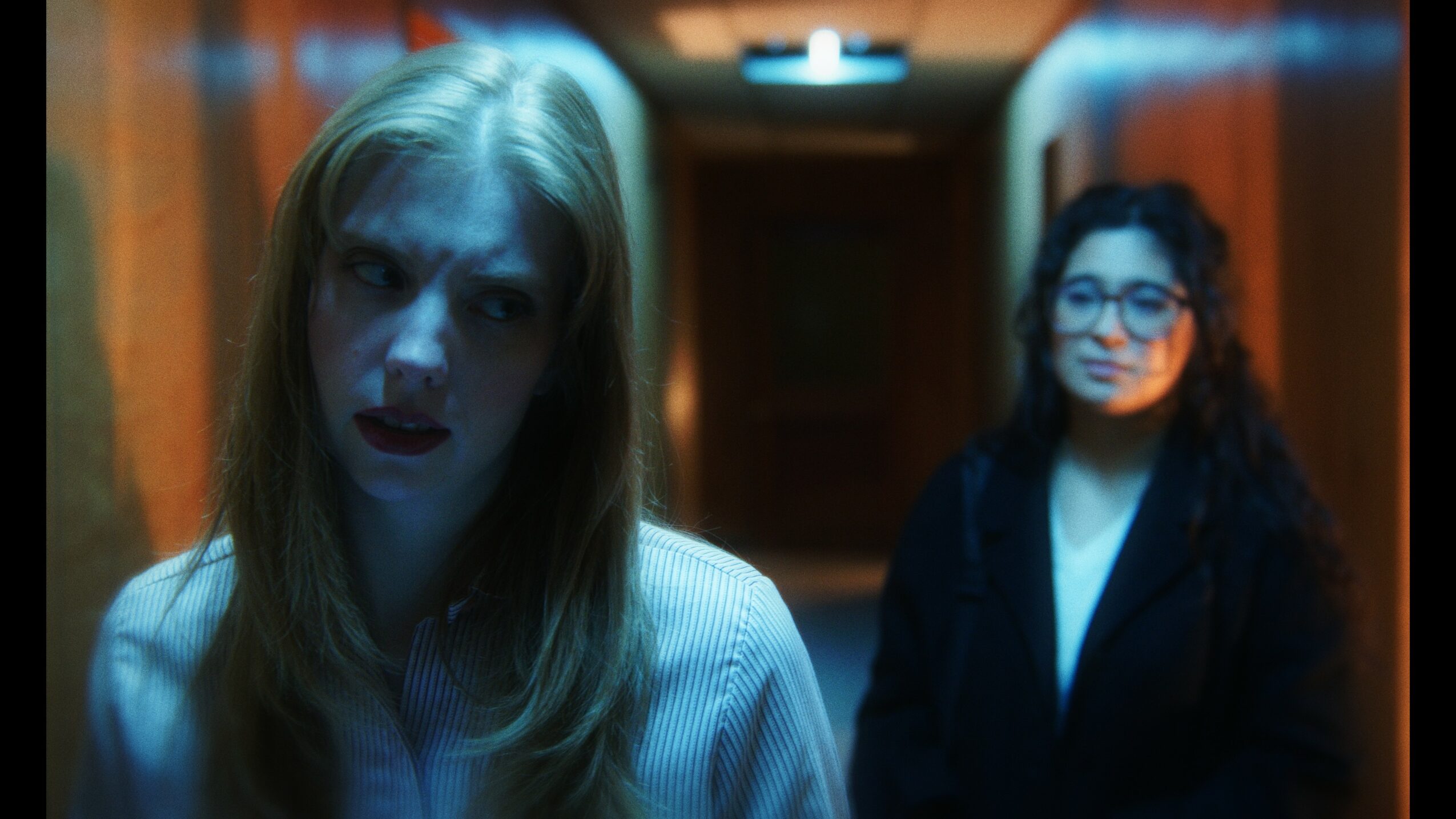 Two women standing in a dimly lit hallway, one in the foreground with light hair and the other in the background with dark hair and glasses.