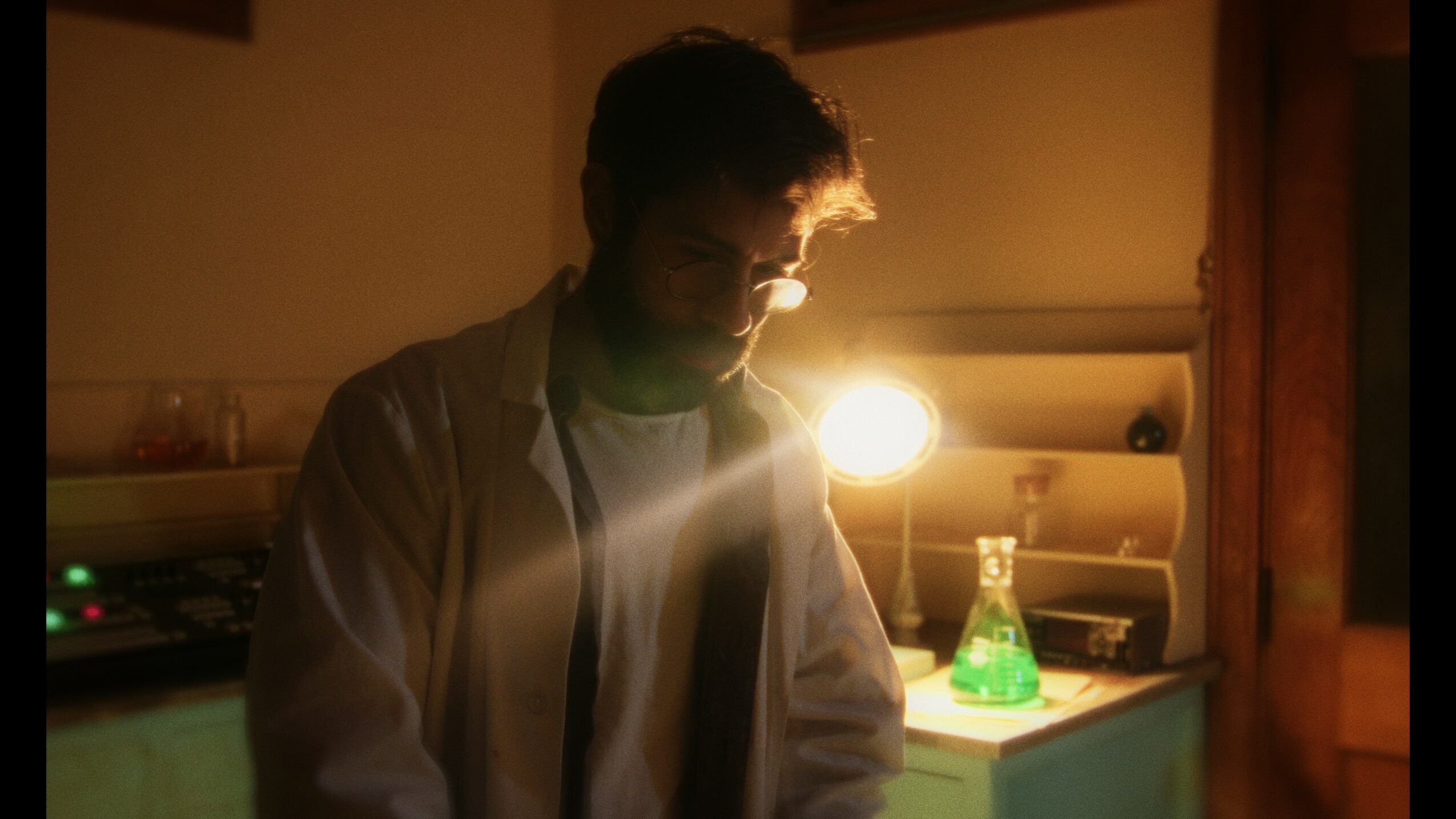 Person with glasses and messy hair in a dimly lit room, illuminated by a bright light behind them, with a lamp and objects on a shelf.
