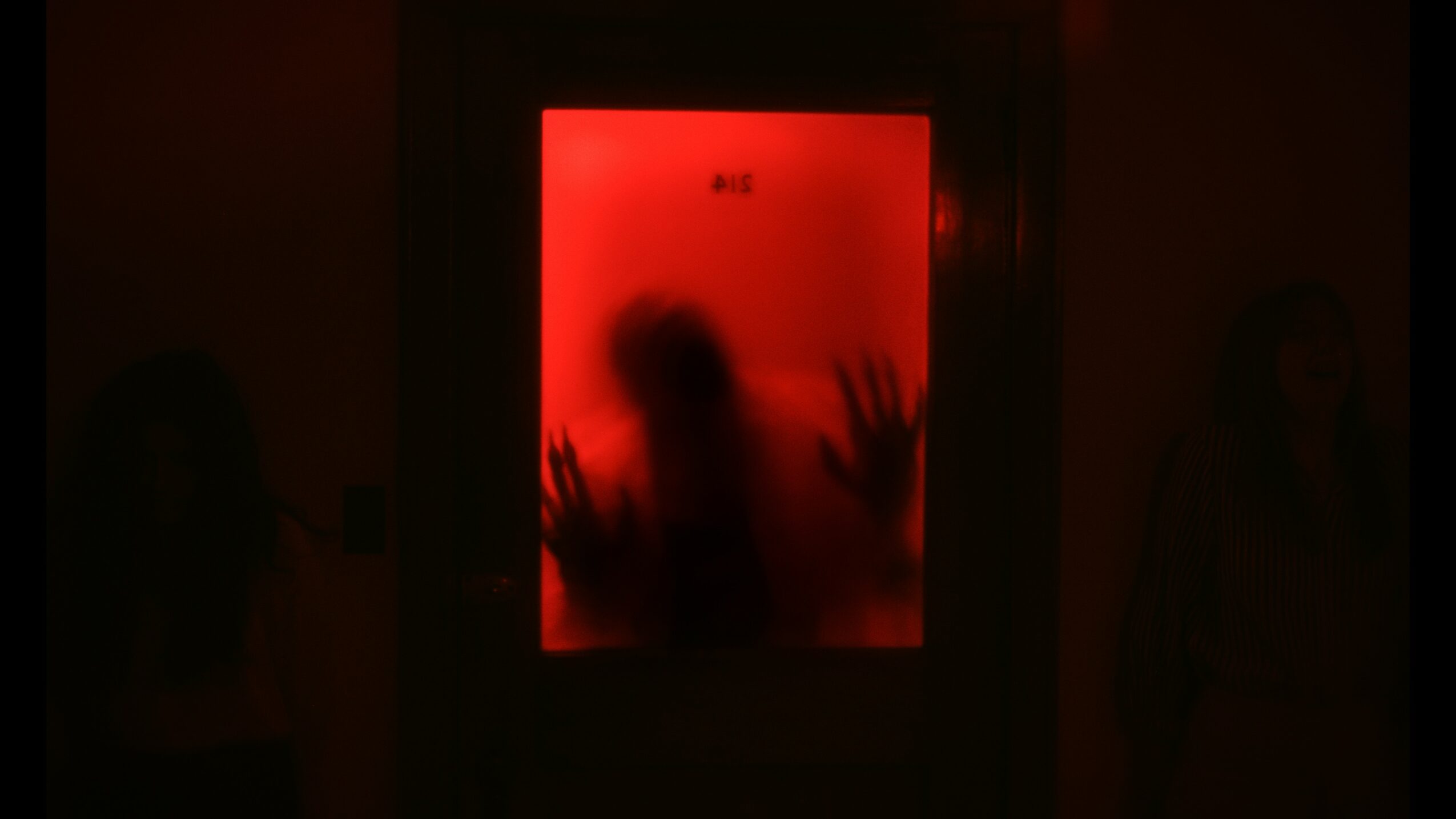 Person pressed against a red, foggy glass door with hands and face visible, surrounded by darkness.