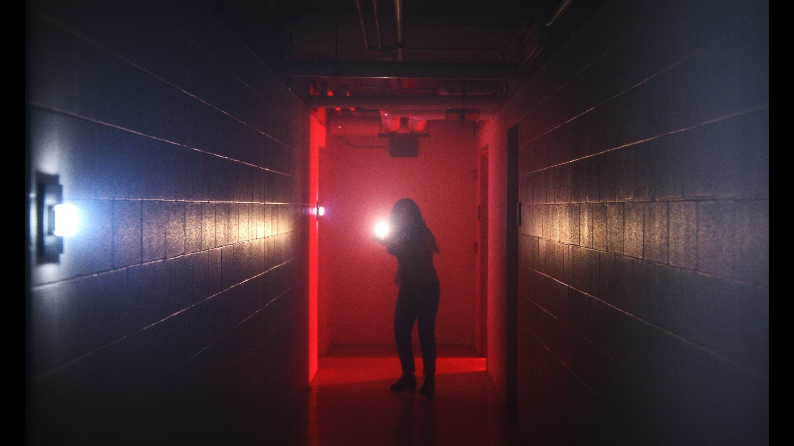 Person standing in a dimly lit corridor with red lighting and laser beams, silhouette visible against the red background.