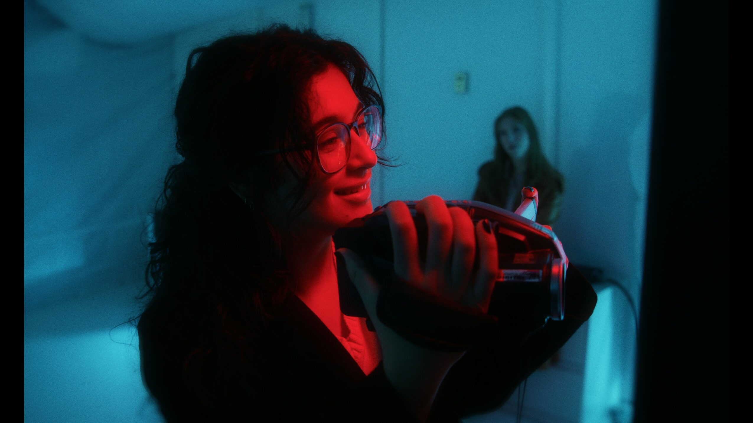 Person with curly hair holding a camera, illuminated by red and blue lighting, with a person in the background.