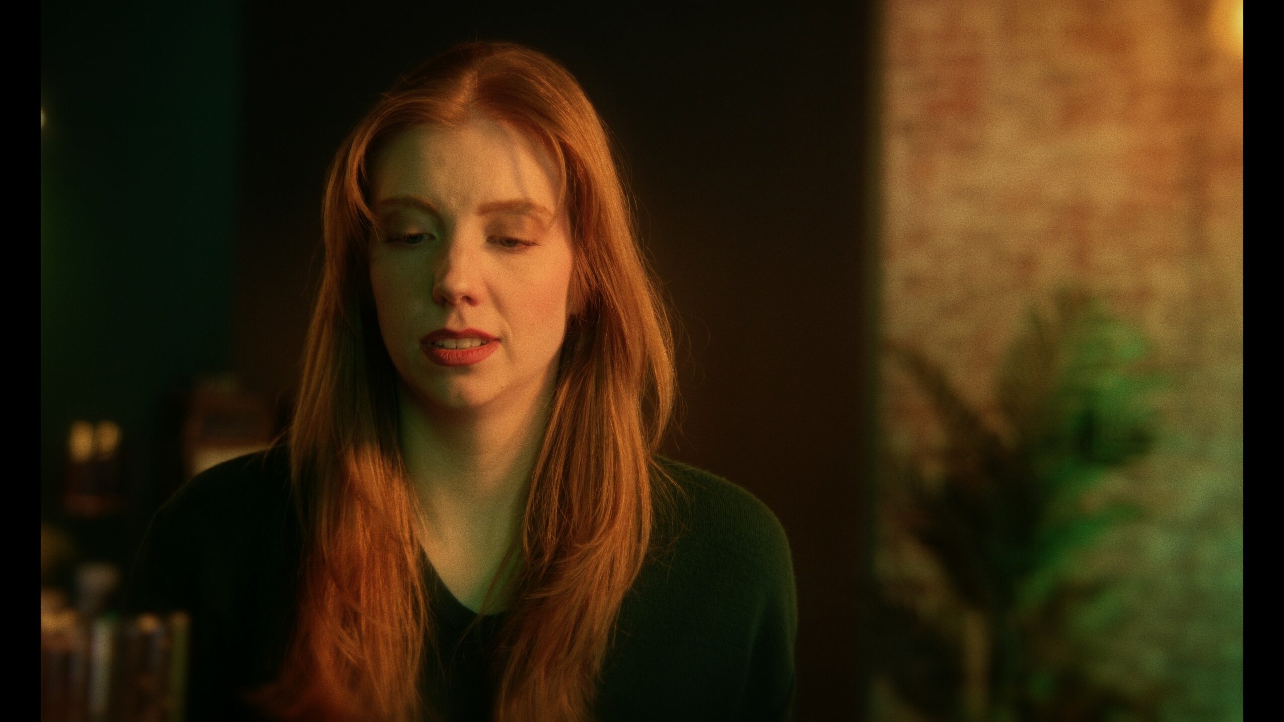 A young woman with long red hair and a black top, looking down, in a dimly lit room.