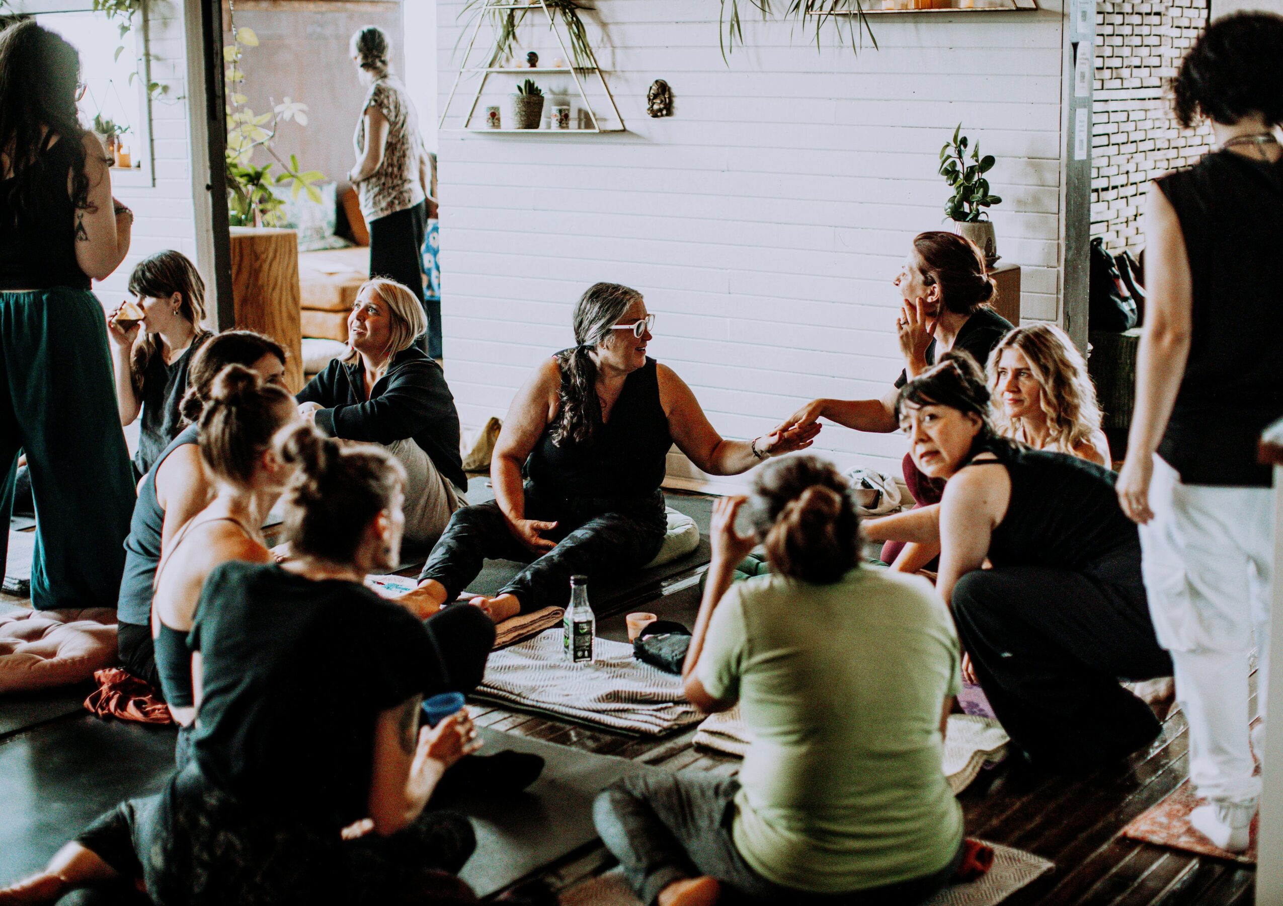 Group of people sitting on the floor in a circle, engaging in conversation indoors with natural light and plants.