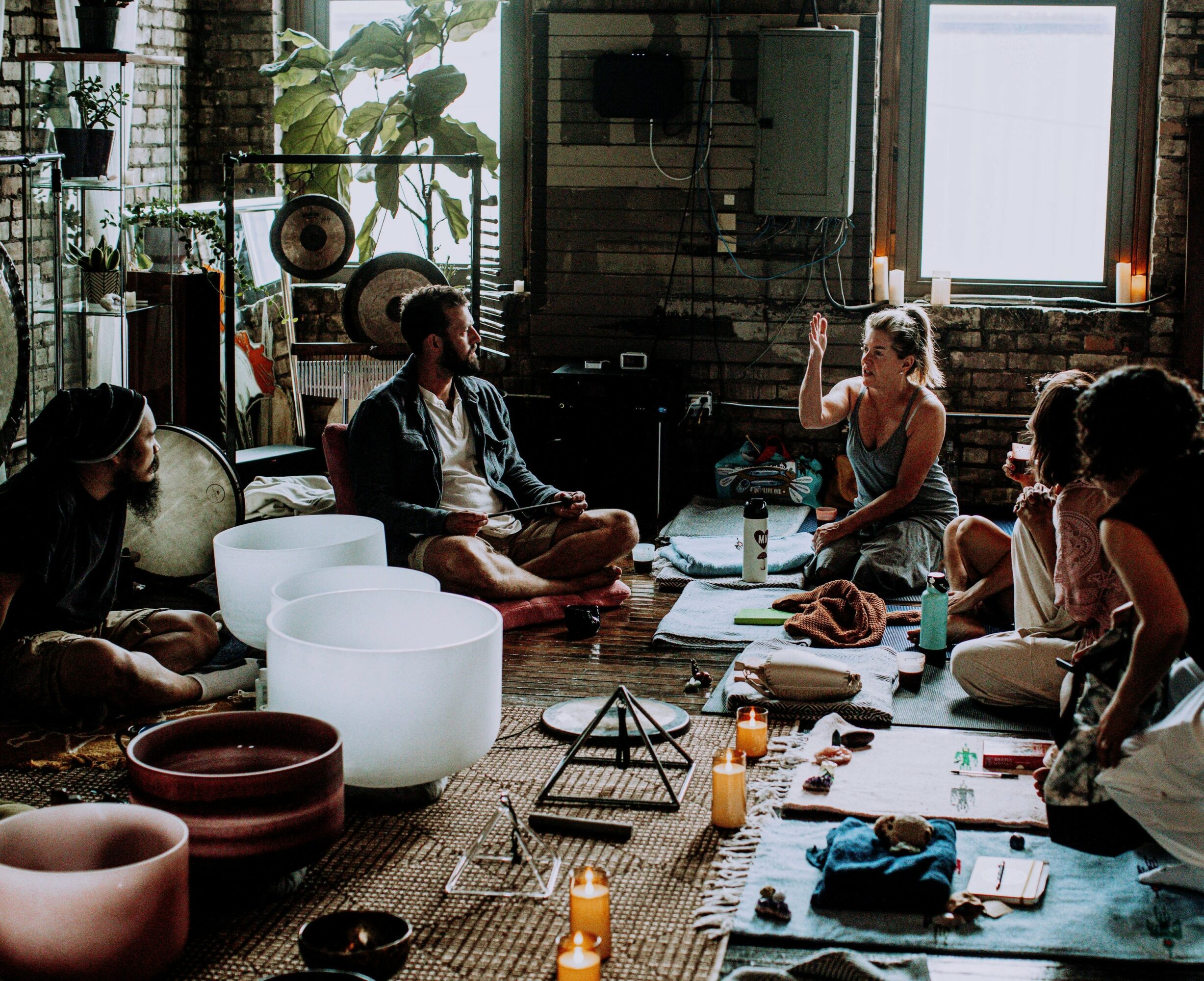 Group of people sitting on the floor in a circle during a meditation or discussion session in a rustic room with large windows.