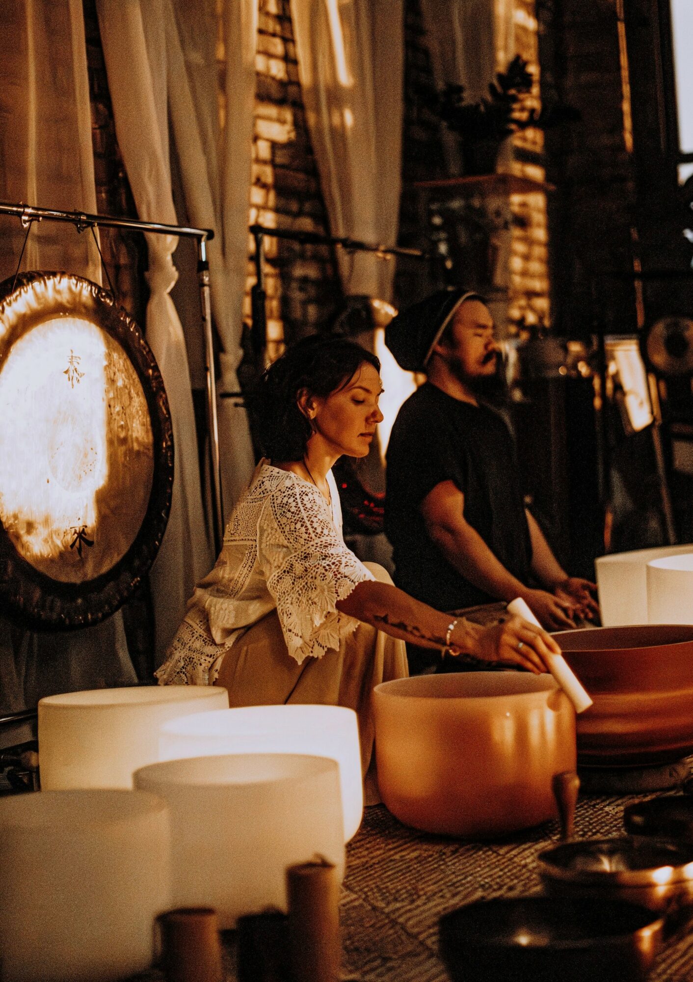 Two people playing large singing bowls indoors with warm lighting and brick walls in the background.