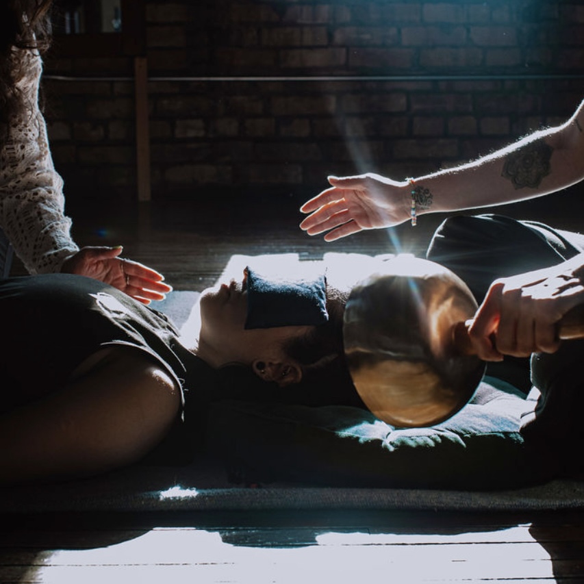 Person lying on the floor with a bowl on their head, surrounded by others' hands, in a dimly lit room.