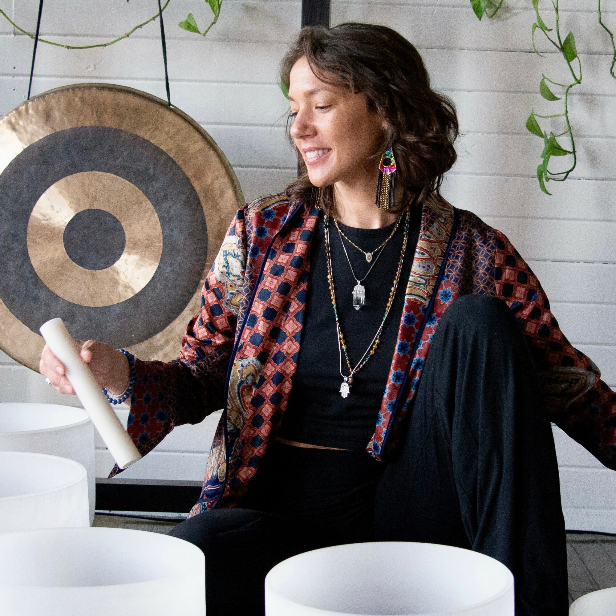 Woman smiling, holding a gong, sitting indoors with plants and white wall background.