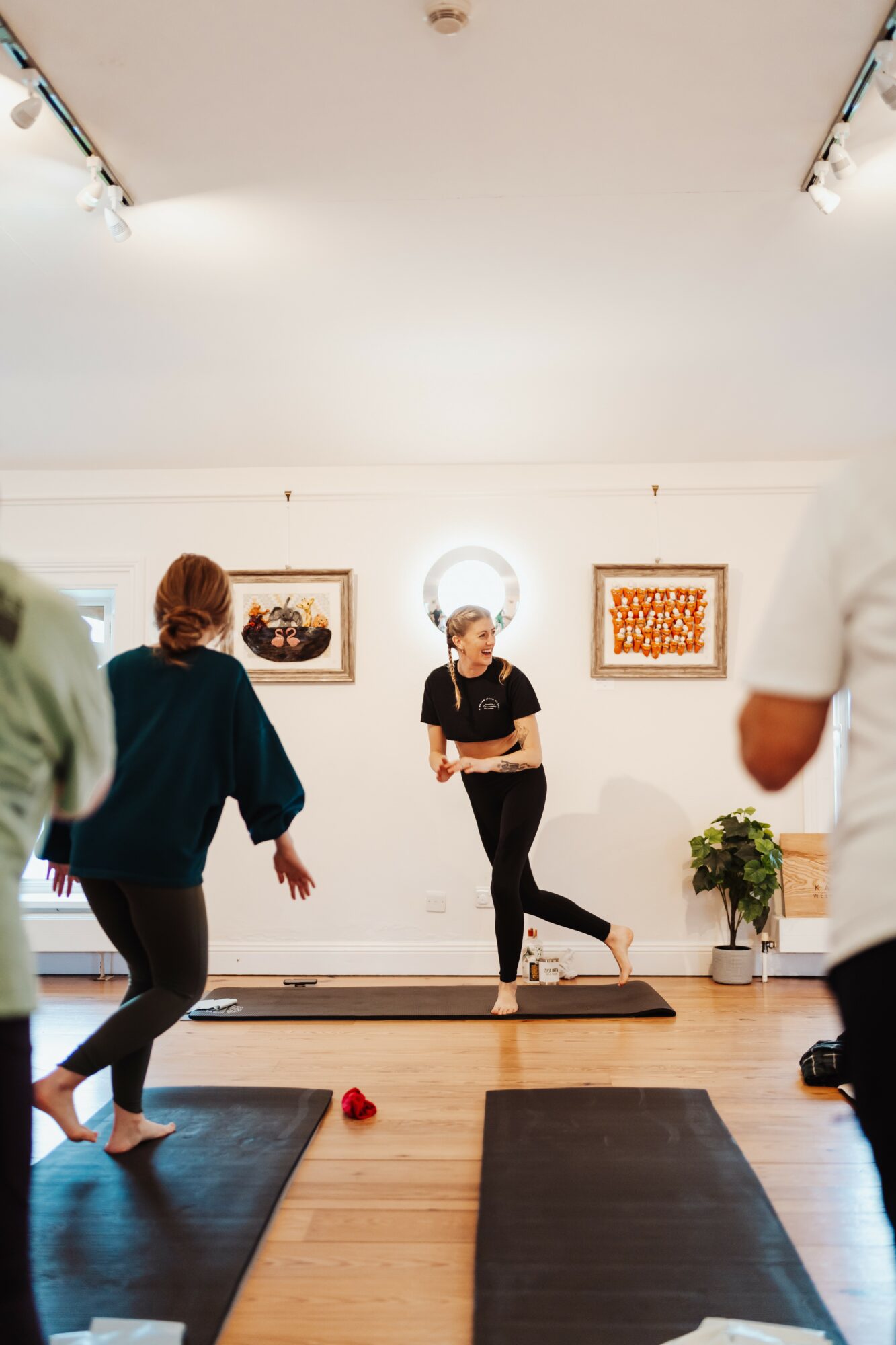 People participating in a fitness class in a studio with artwork on the wall, a woman leading, and mats on the floor.