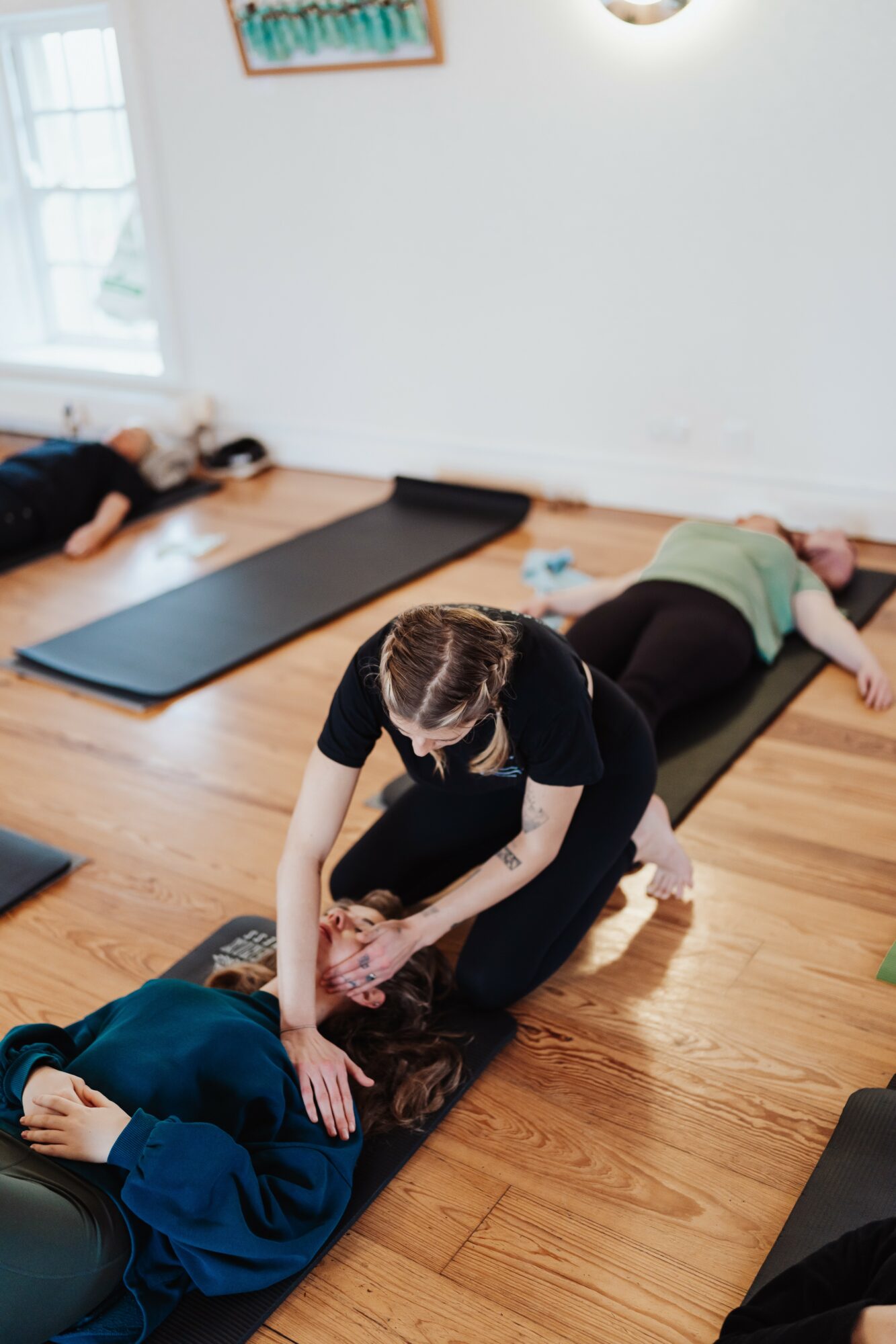 Instructor assisting a person during a yoga class in a bright room with wooden floors and mats.
