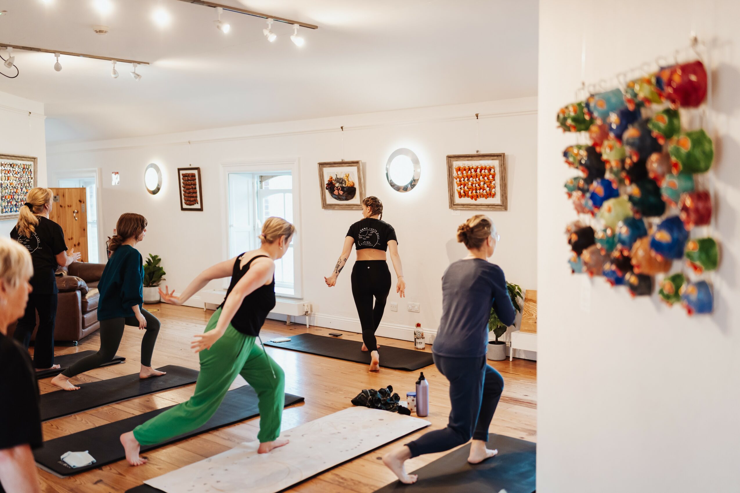Group of people practicing yoga in a bright room with artwork on walls, mats on floor, and a colorful wall hanging.