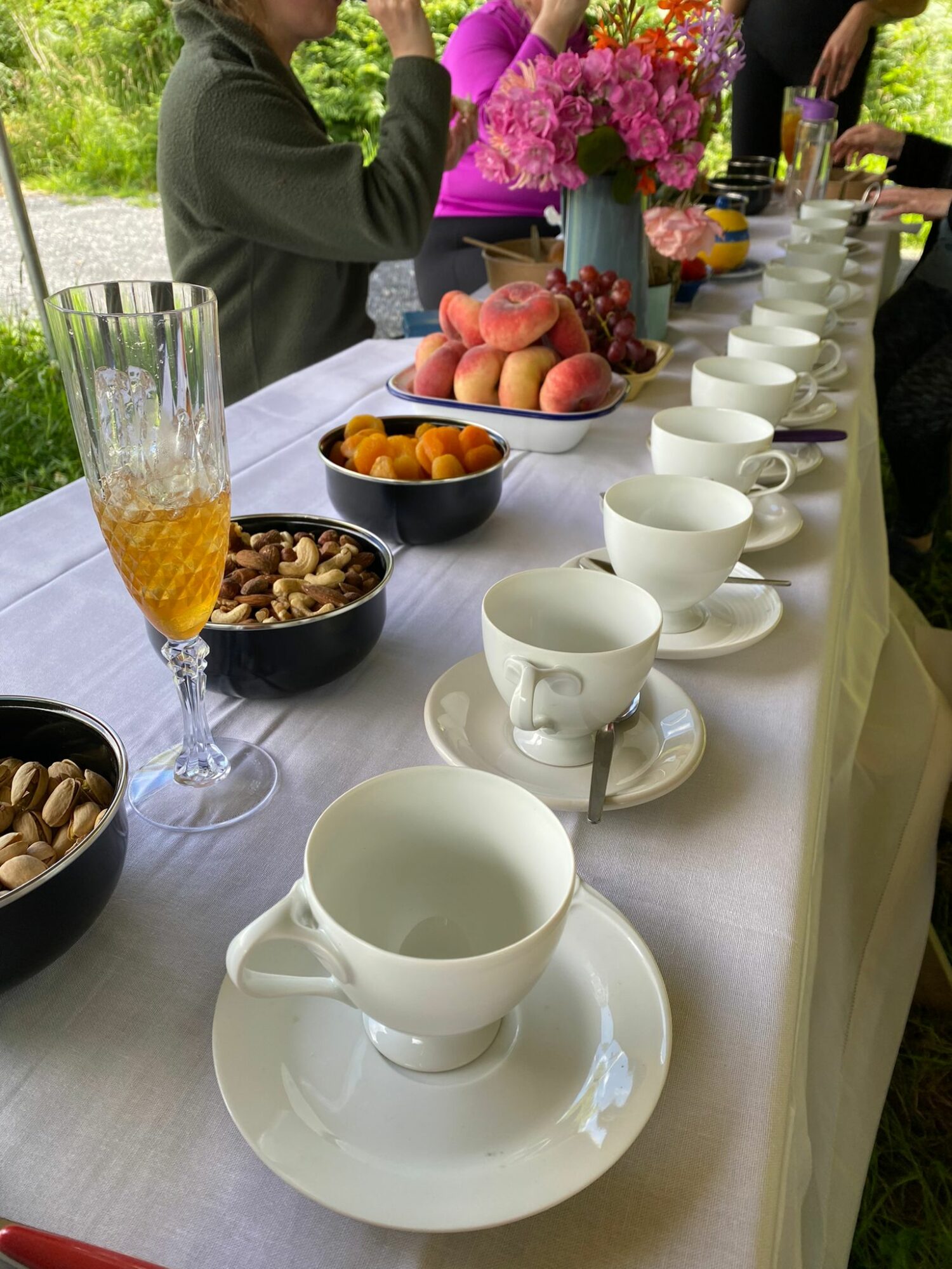 Table set outdoors with teacups, bowls of snacks, a glass of orange drink, and a floral centerpiece, with people in the background.