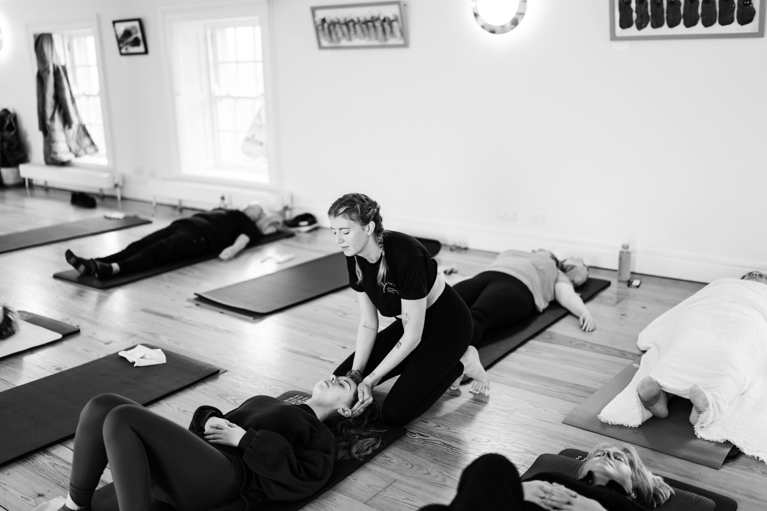 A woman provides first aid to a person lying on the floor in a room with other people, mats, and windows.