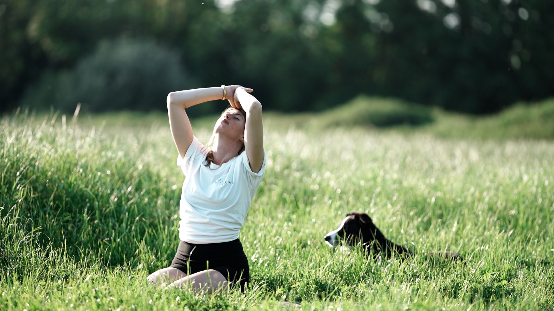 Person kneeling on grass with hands on head outdoors, dog lying nearby, green trees in background