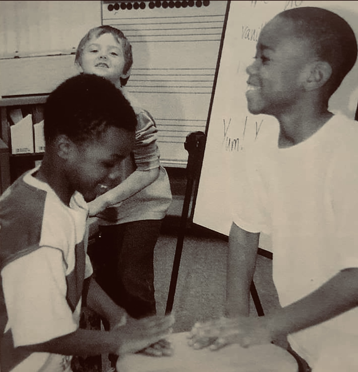 Three children in a classroom, two are shaking hands, one stands in the background near a whiteboard.