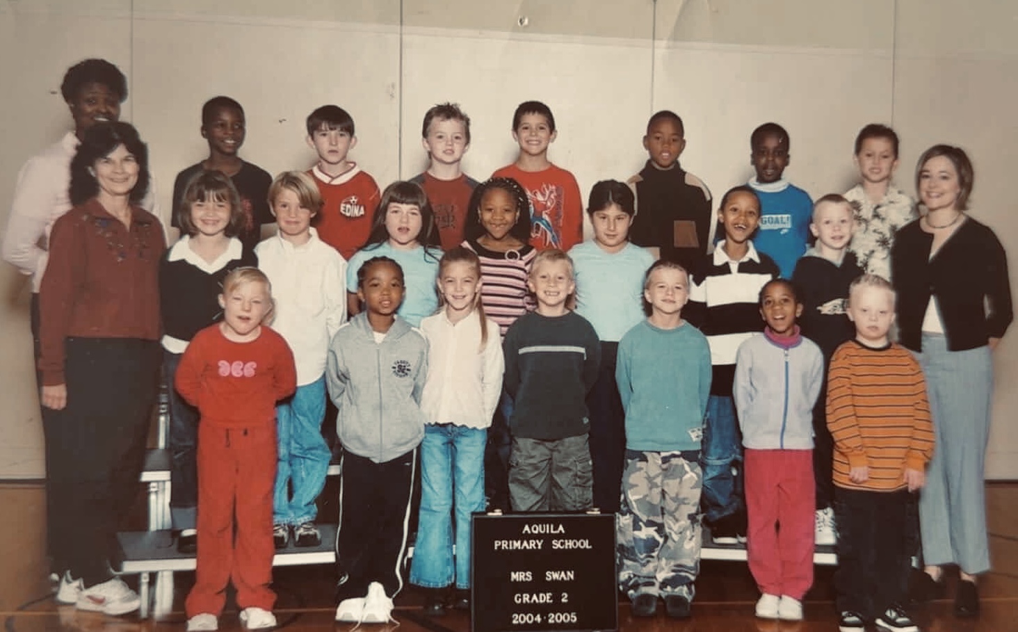 Group of children and two adults in a classroom, with a blackboard displaying names and years, smiling for a photo.