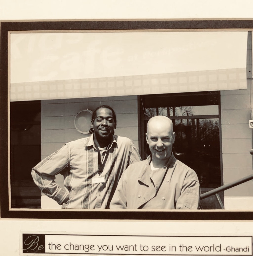 Two men standing outside a house, smiling, with a window and a door behind them.