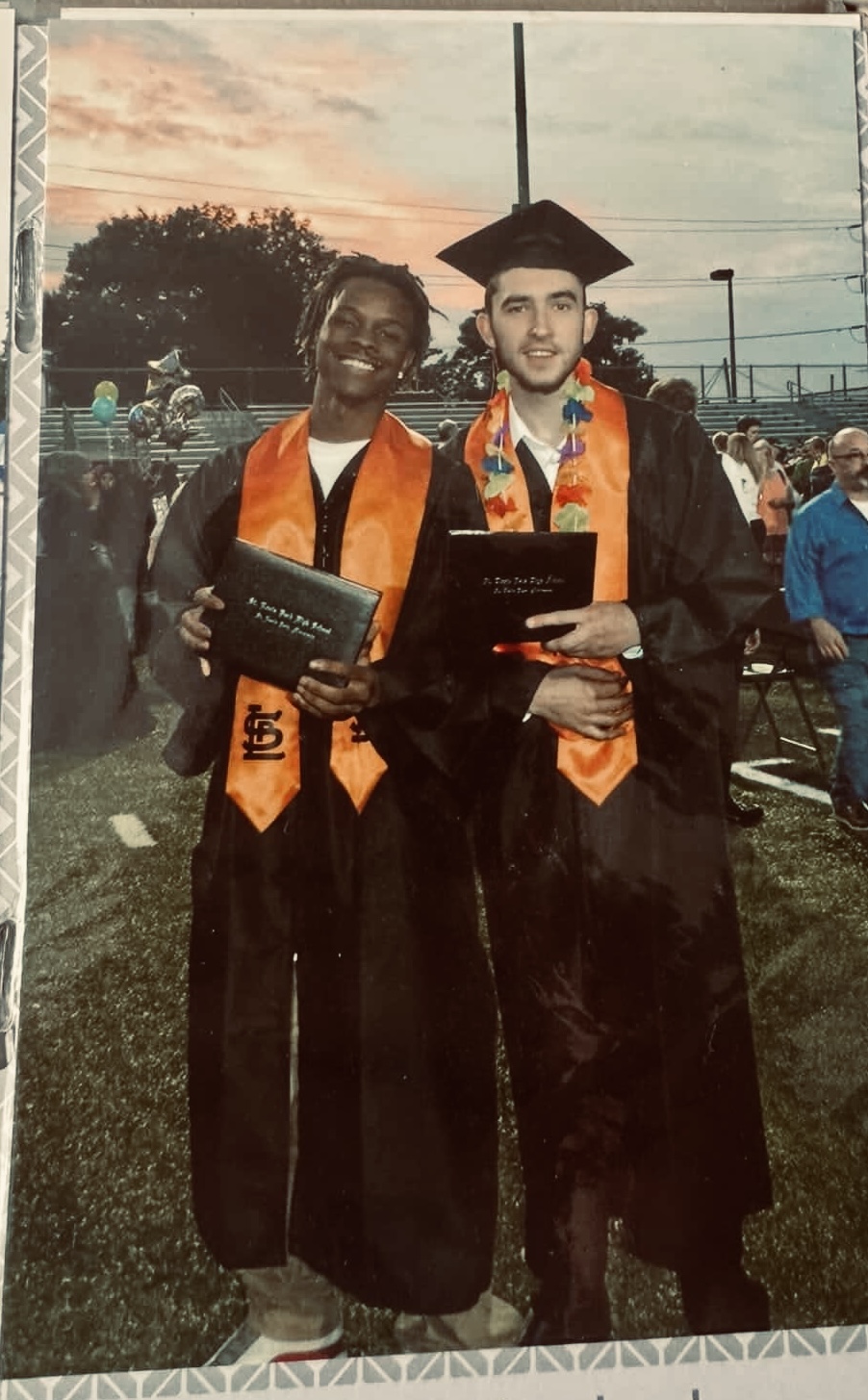 Two graduates in caps and gowns holding diplomas, smiling outdoors during graduation ceremony, with other people and trees in background.