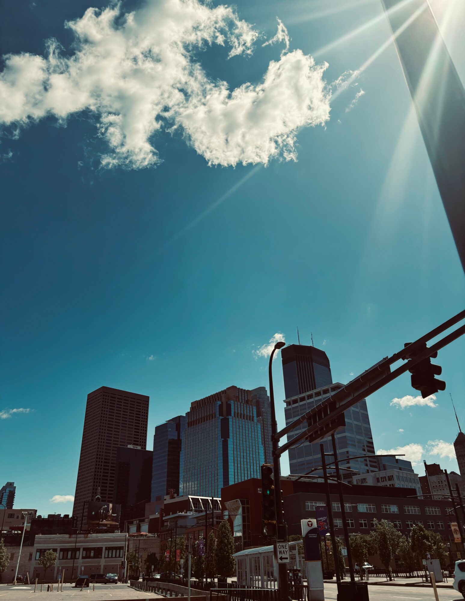 City skyline with tall buildings under a bright blue sky and sunlight, with traffic lights in foreground.