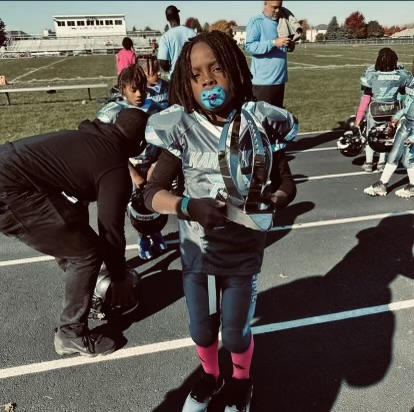 Child holding a trophy on a sports field, wearing a silver sports jersey, face mask, and pink socks, with other children and adults in the background.