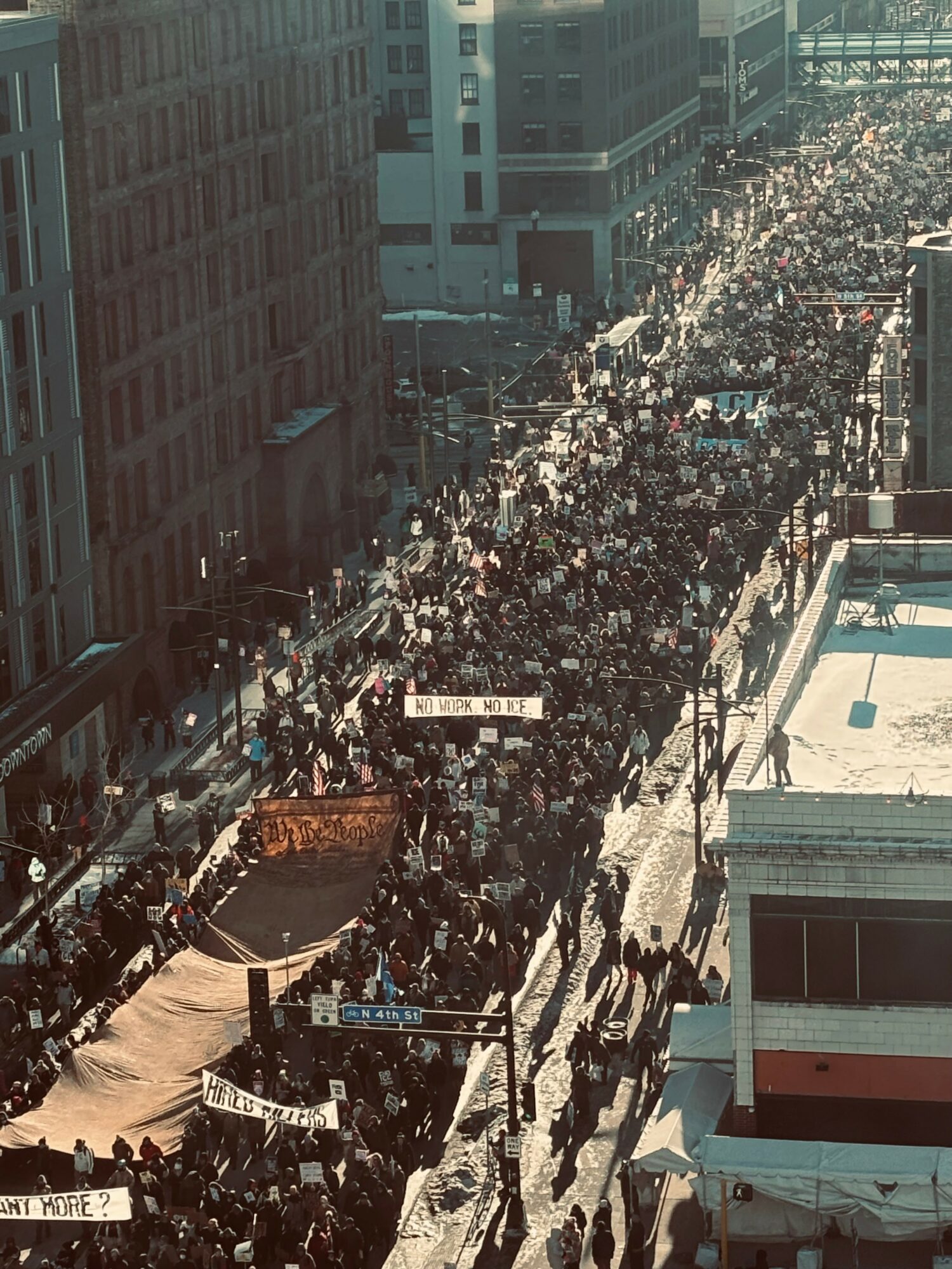 Crowd of people marching on a city street with tall buildings, some holding banners and signs, sunlight casting shadows.