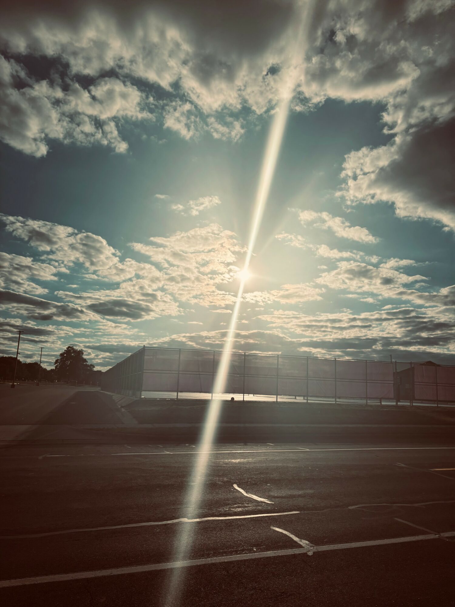 Sunlight streams through clouds in a partly cloudy sky over an empty parking lot with a fence in the background.