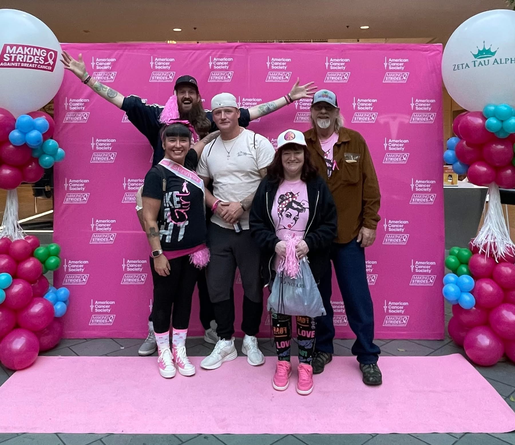Group of five people standing on pink carpet in front of pink backdrop with balloons on sides, smiling and posing for photo.
