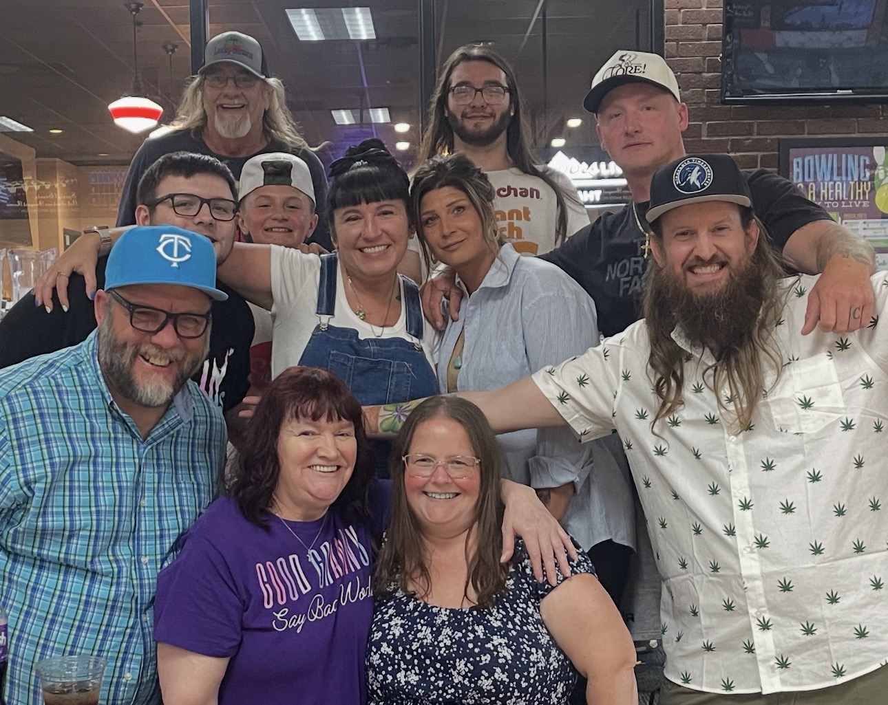 Group of ten people smiling together indoors, some wearing hats and glasses, with a brick wall and TV screen in background.