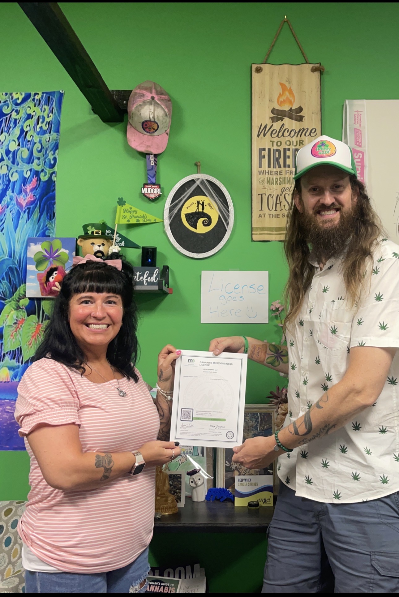 Two people smiling and holding a certificate in a colorful room with various decorations and signs.