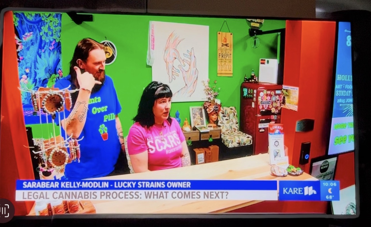 Two women sit at a table in a colorful room with a green screen background, a drawing on the wall, and various items on the table.
