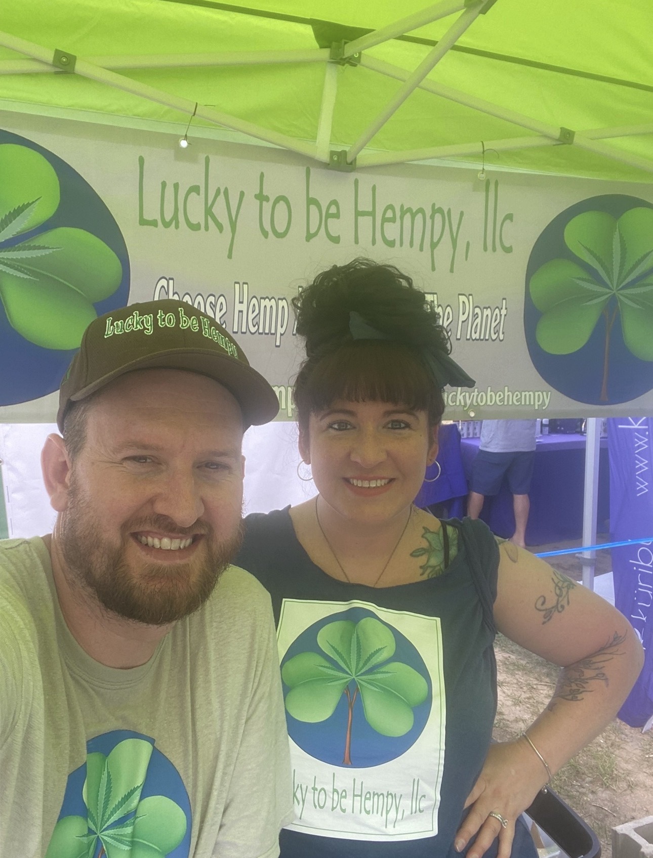 Two people smiling in front of a green tent with a sign and logo for Lucky to be Hempy, Inc.