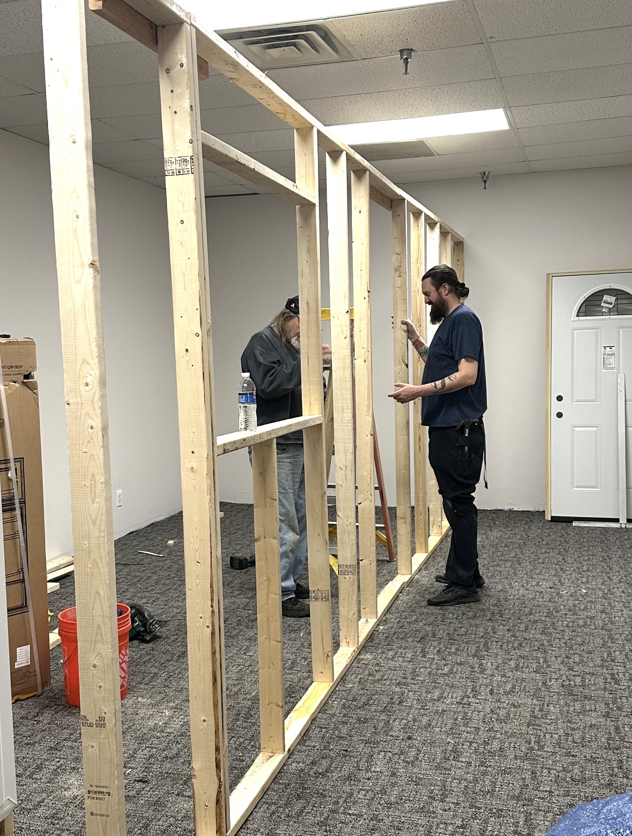Two people working on a wooden frame in an indoor space, with a door and a vending machine in the background.