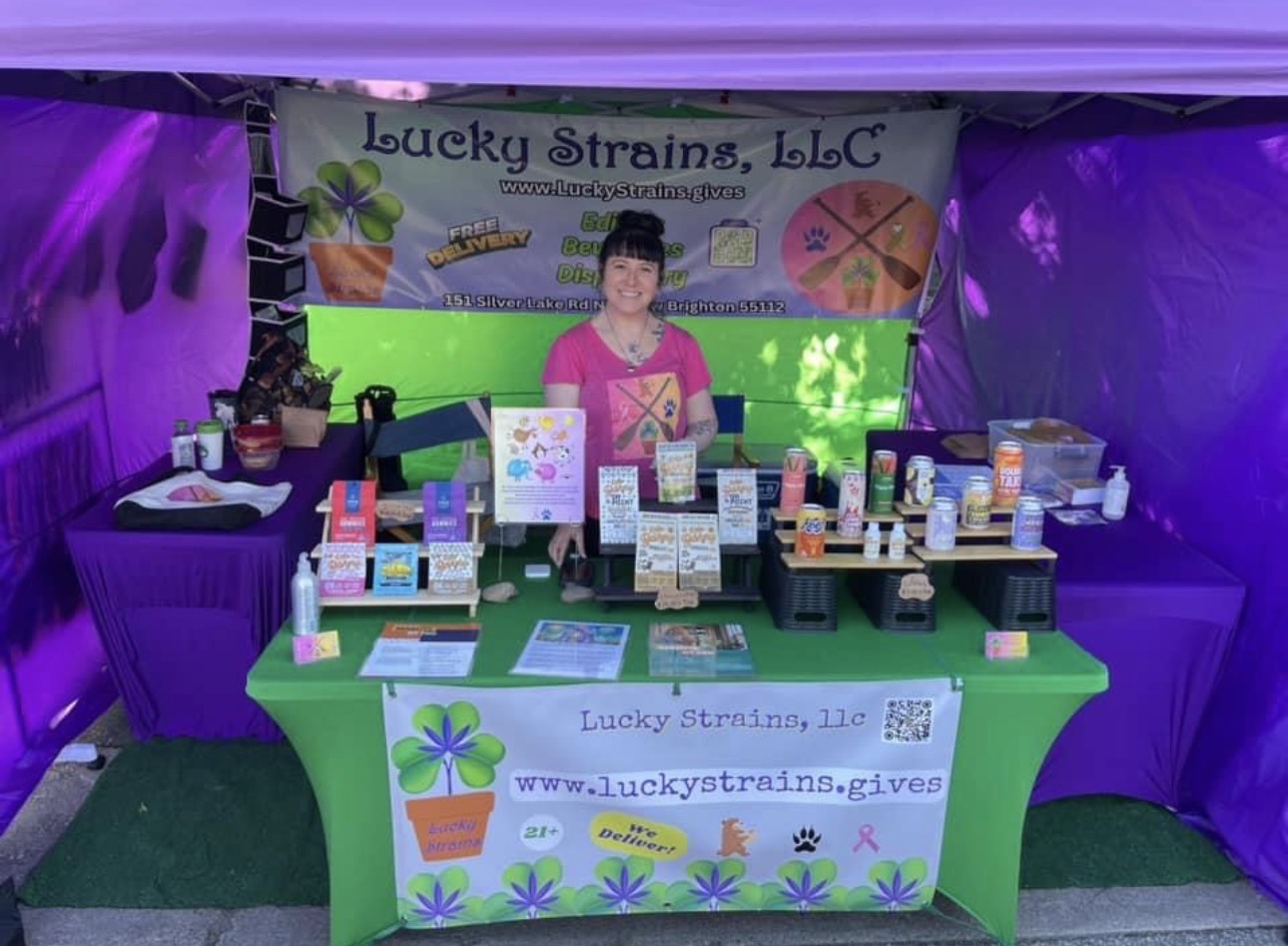 Woman standing behind a table with health products at Lucky Strains, LLC booth, purple tent background, green table cover, banner, and products displayed.