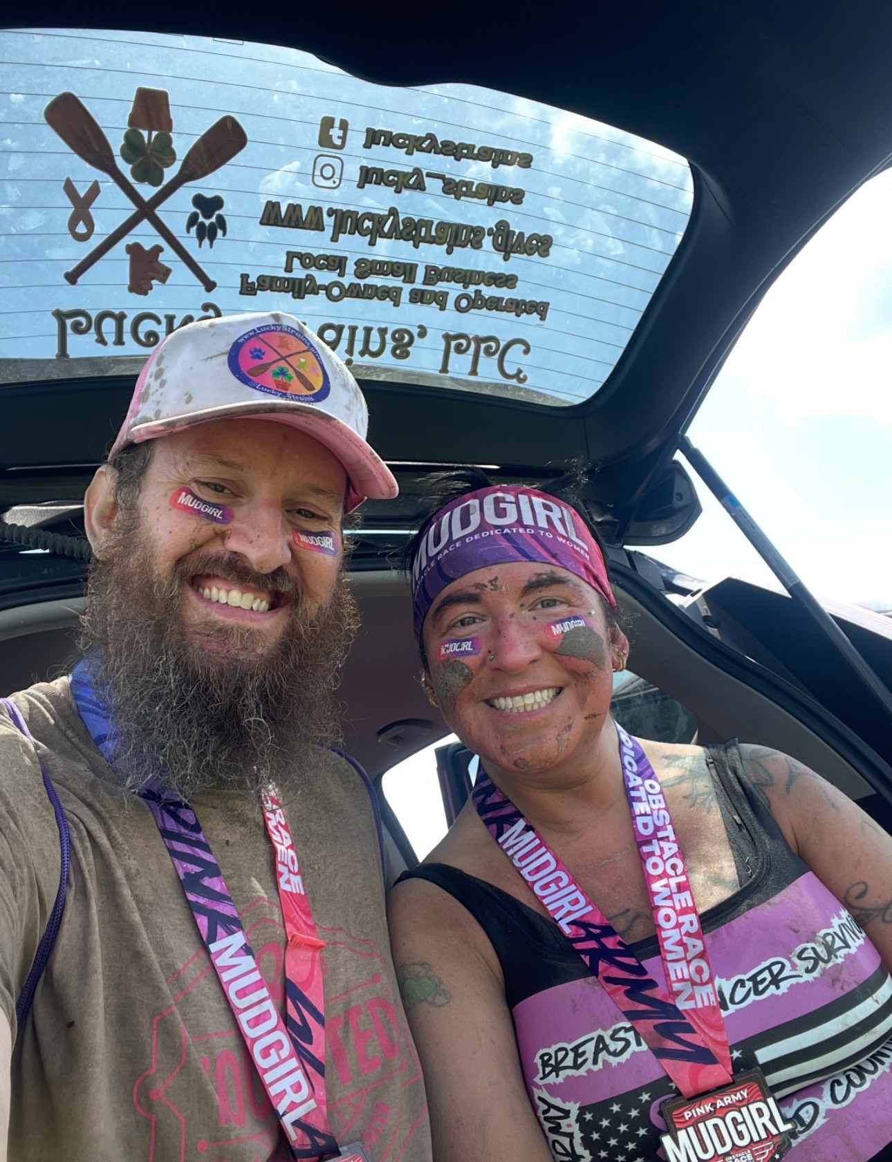 Two smiling people with face paint and medals inside a vehicle, with a stickered window behind them.