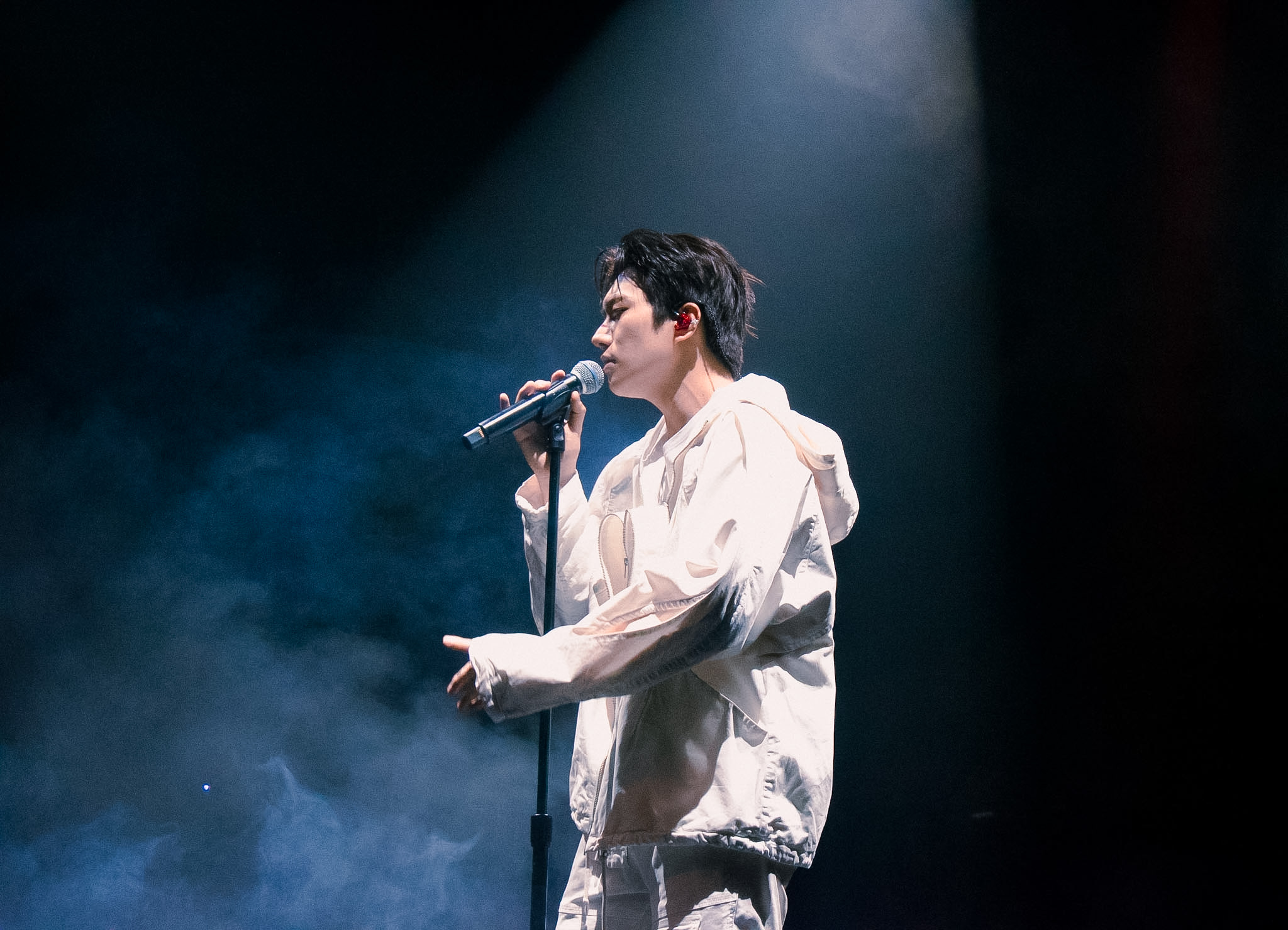 Person singing into a microphone on stage with dark background and spotlight, wearing light-colored clothing.