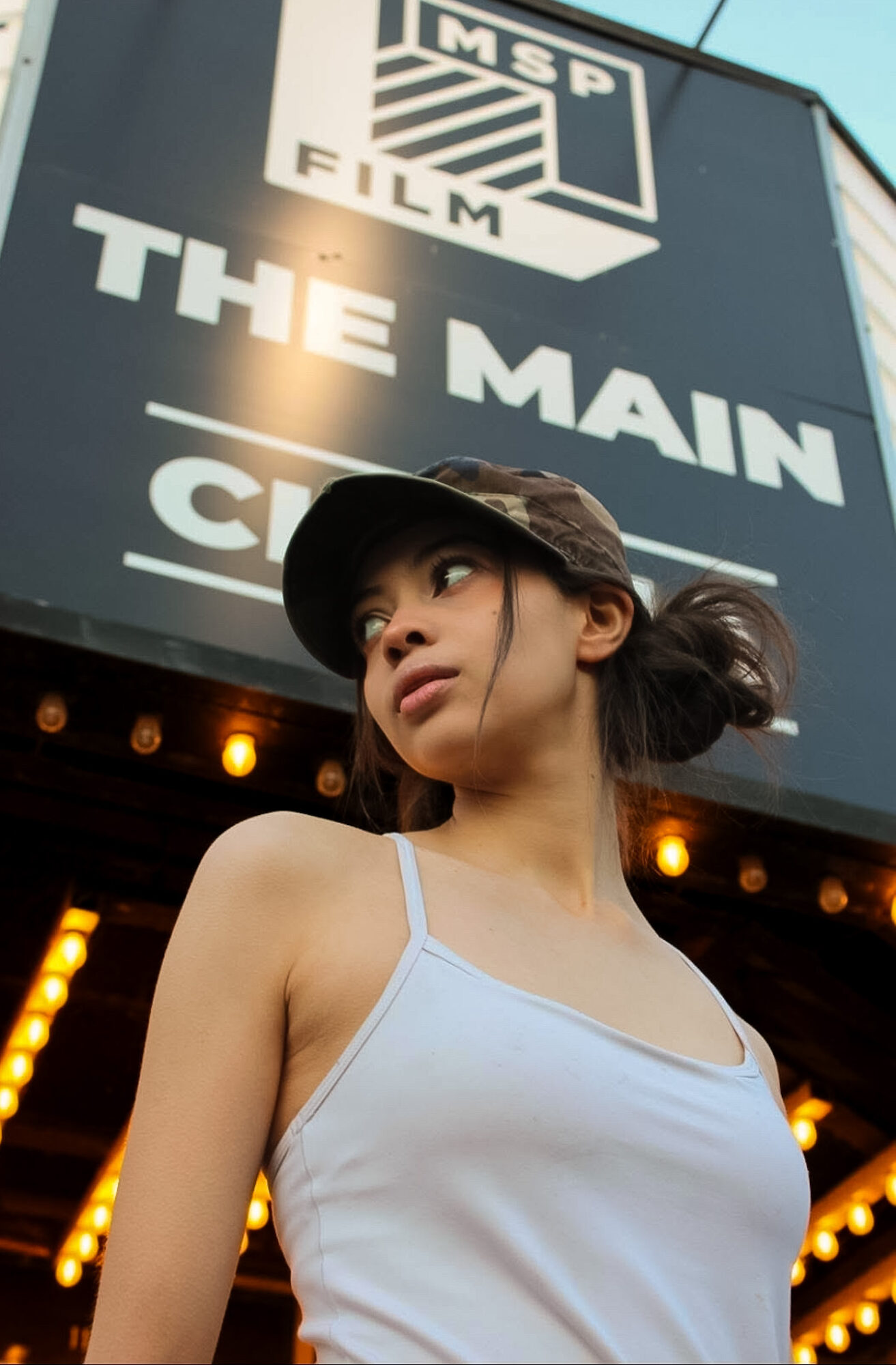 Young woman with dark hair in a ponytail, wearing a white tank top and camouflage cap, looking to the side outdoors.