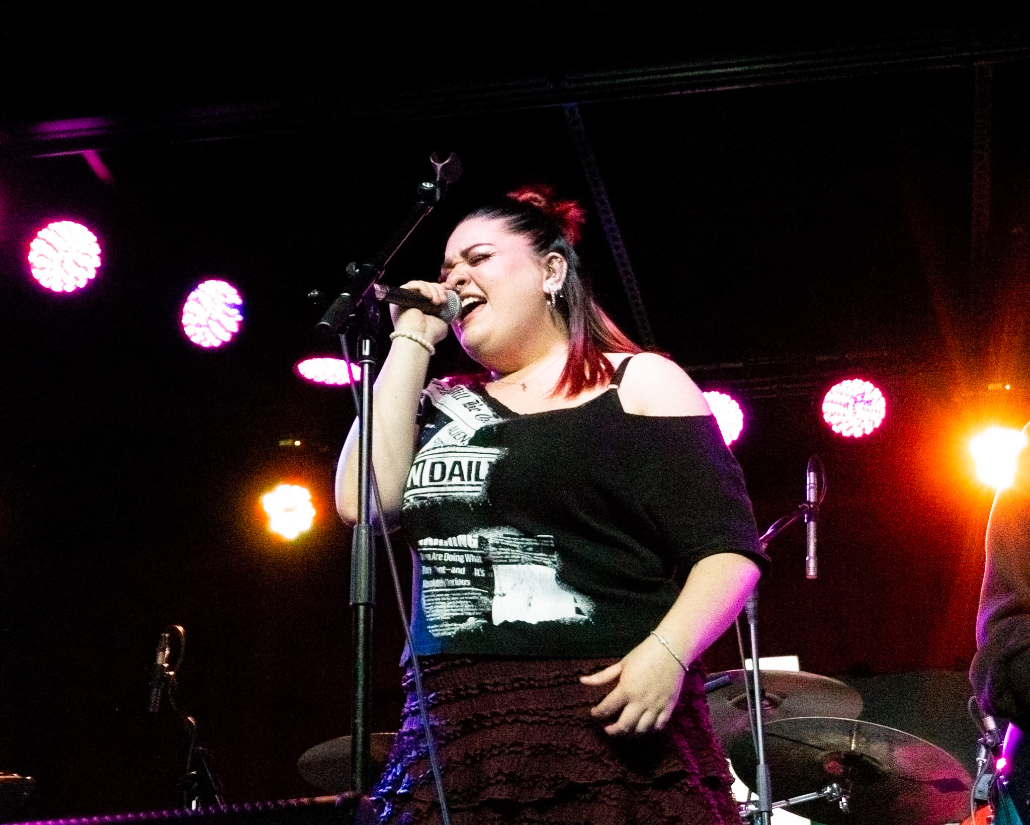 Young woman singing into a microphone on stage with colorful lights behind her.