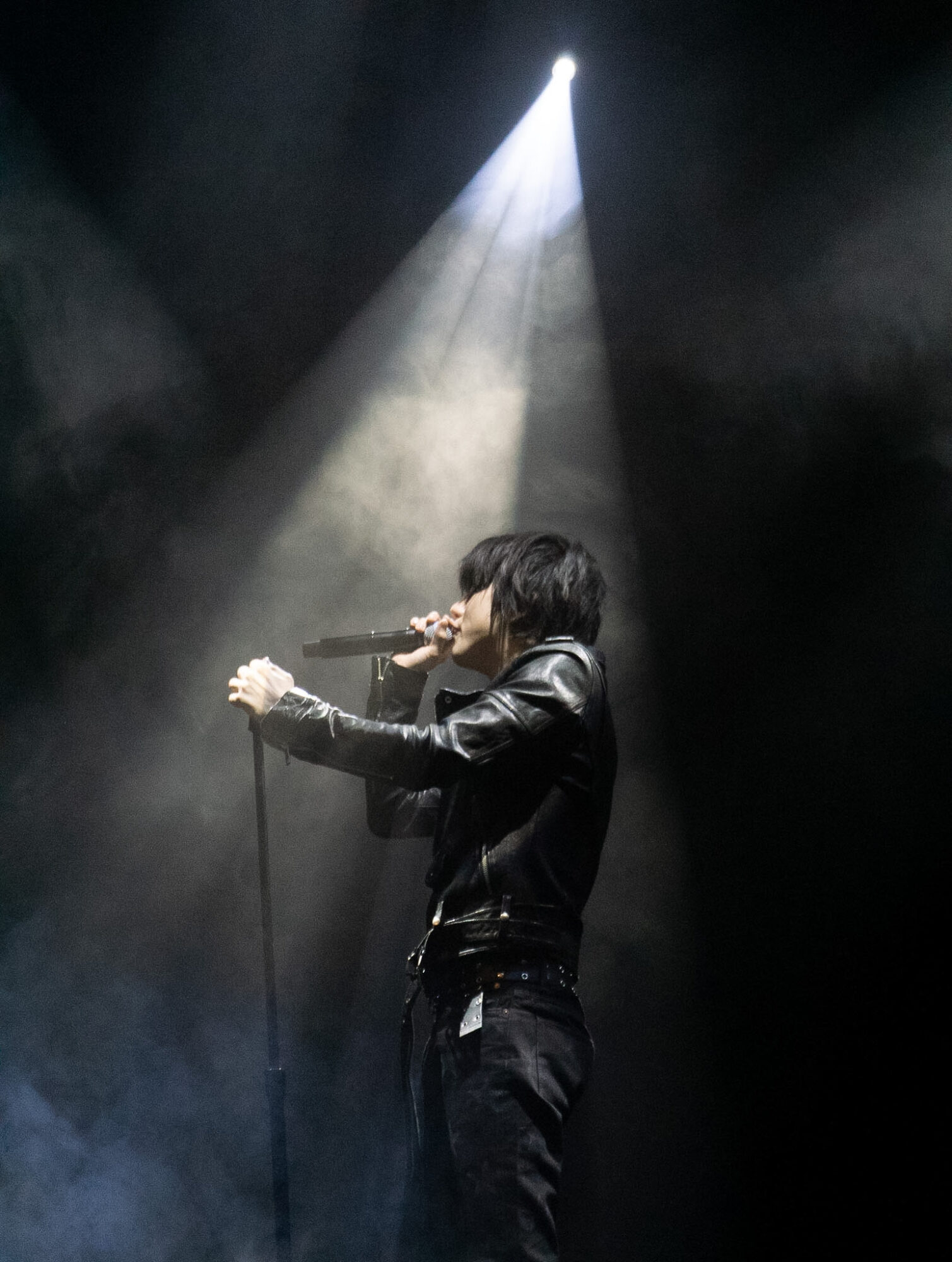 Person singing into microphone on stage with spotlight shining down, dark background, wearing dark clothing.