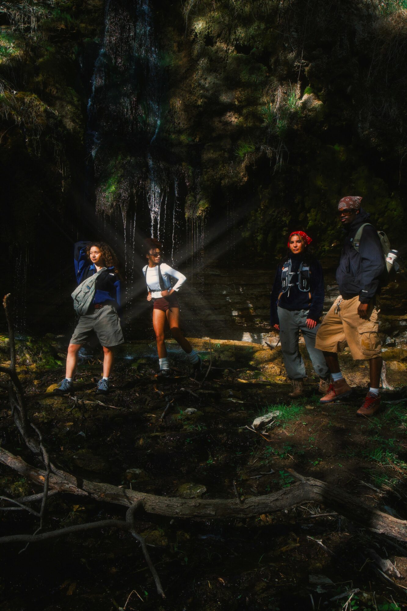 Four people stand outdoors near a waterfall, surrounded by trees and rocks, with sunlight filtering through the foliage.