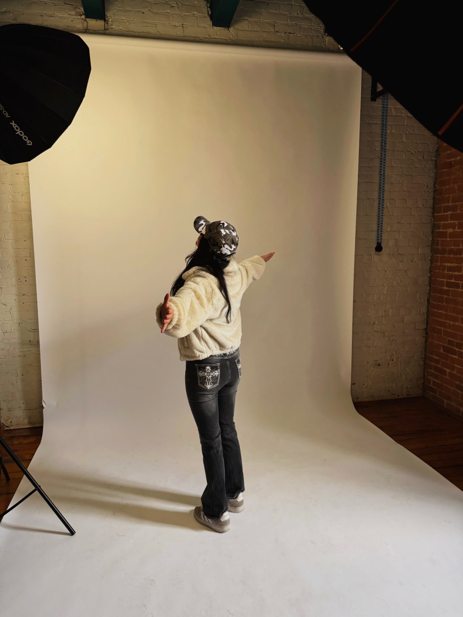 Person standing in front of a white backdrop, pointing to the right, wearing a shiny hat, white jacket, and jeans.