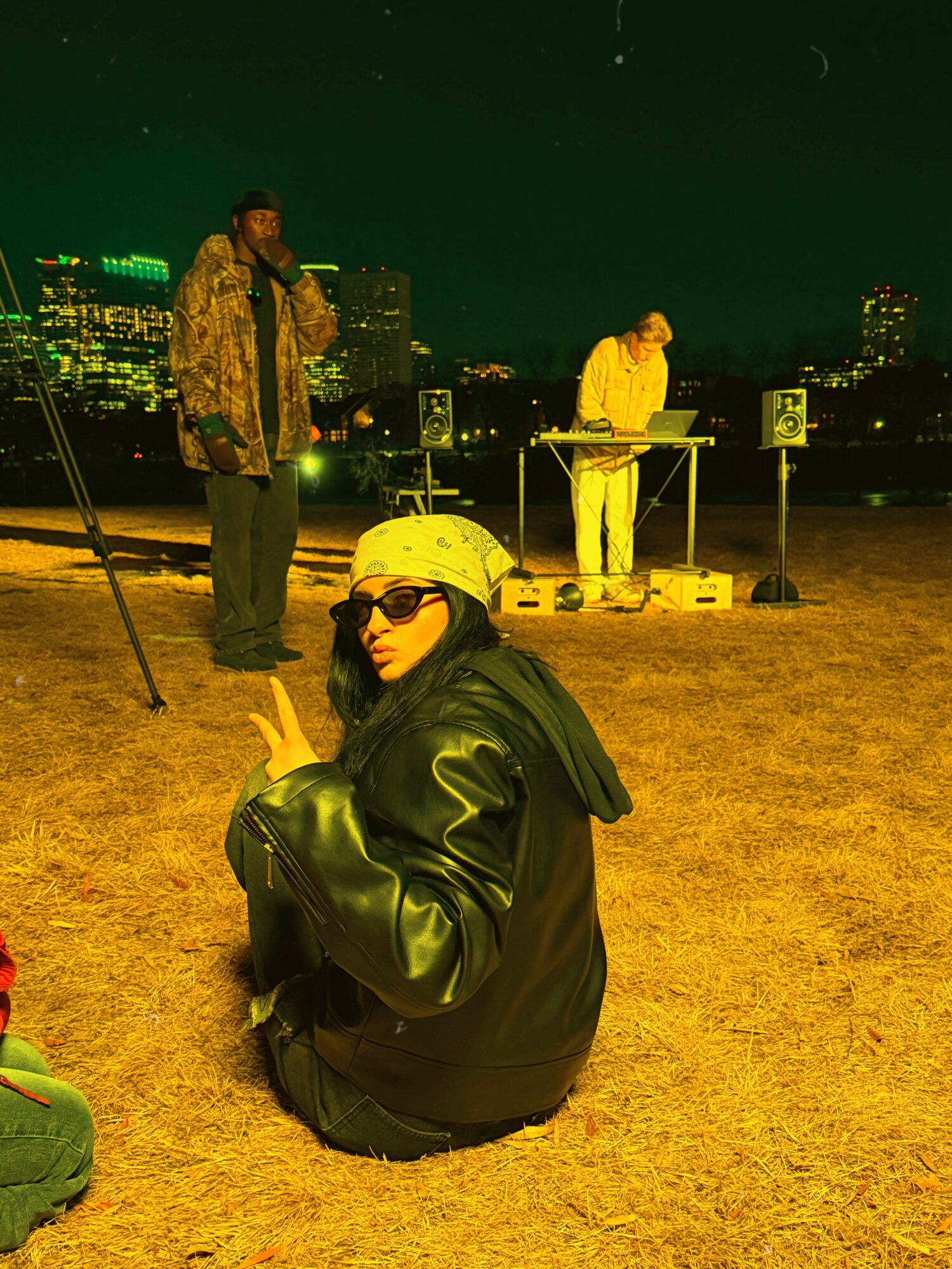 Person sitting on ground wearing sunglasses and a black jacket, with a city skyline at night in the background.