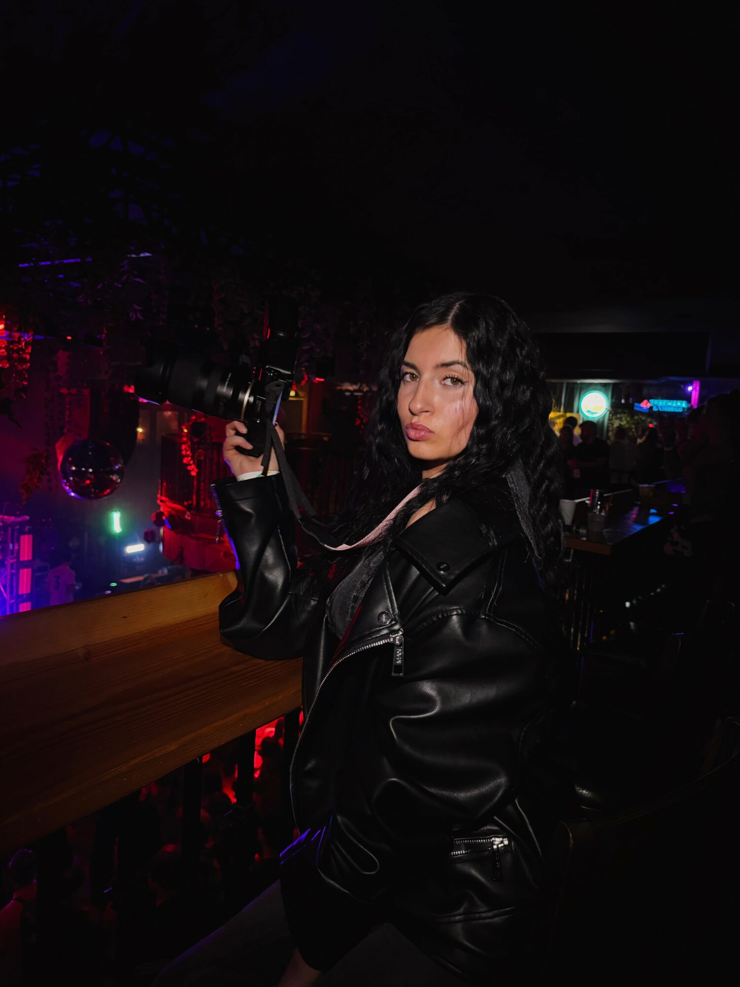 Woman with dark curly hair in a black leather jacket holding a gun in a dimly lit nightclub.